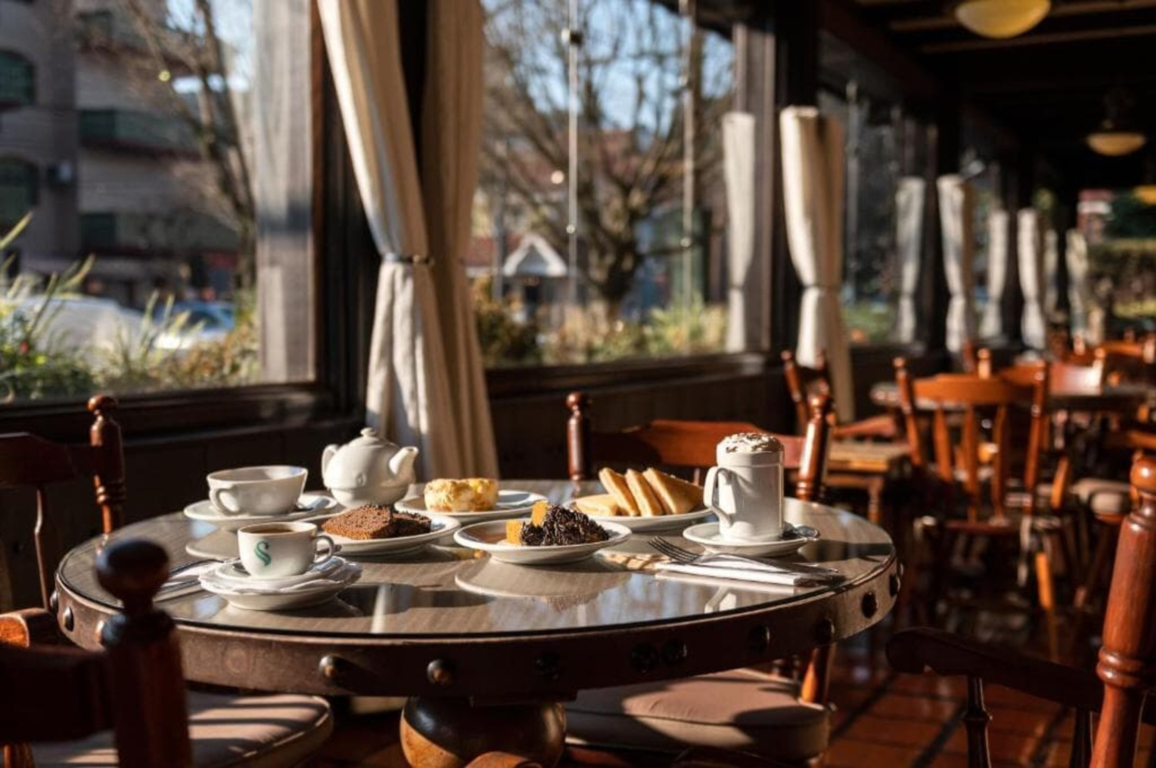 Una mesa redonda con una tetera, tazas y pasteles en una cafetería con grandes ventanales y sillas de madera.