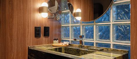 A bathroom with a wooden wall, a large mirror, two wall-mounted lights, a sink with a marble countertop, and a window with blue-tinted glass.