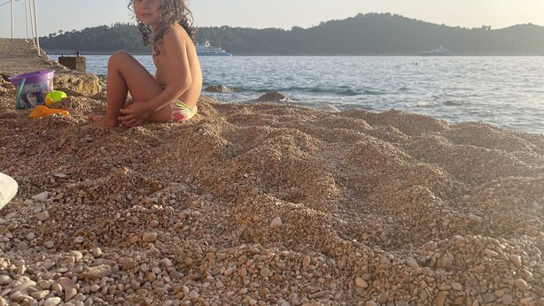 A child sitting on a pebbled beach with a clear view of the sea and distant hills. - Rilati Old Town Palace (Dubrovnik)