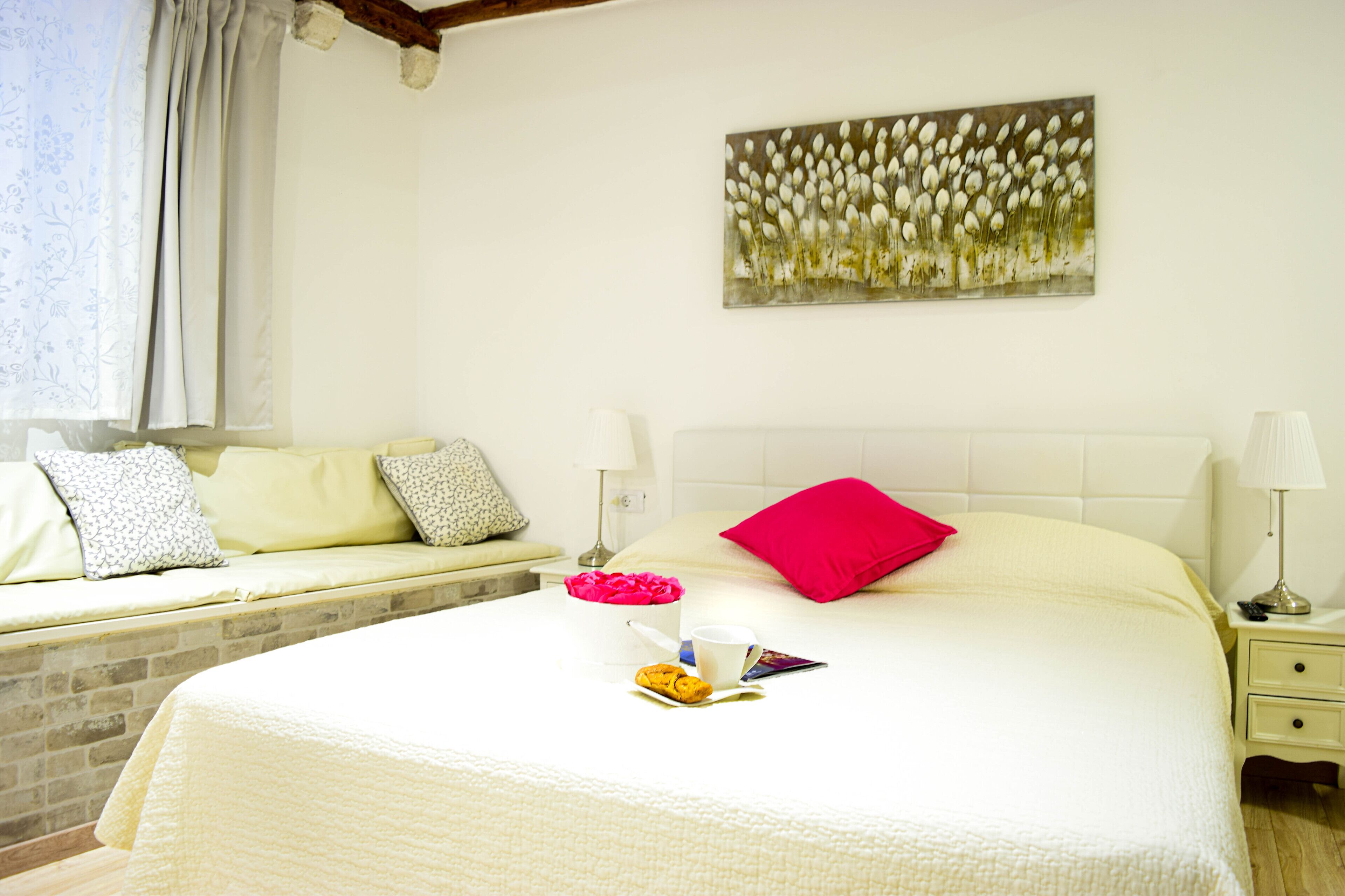 A neatly made bed with a white comforter, a red pillow, and a small bedside table with a lamp.