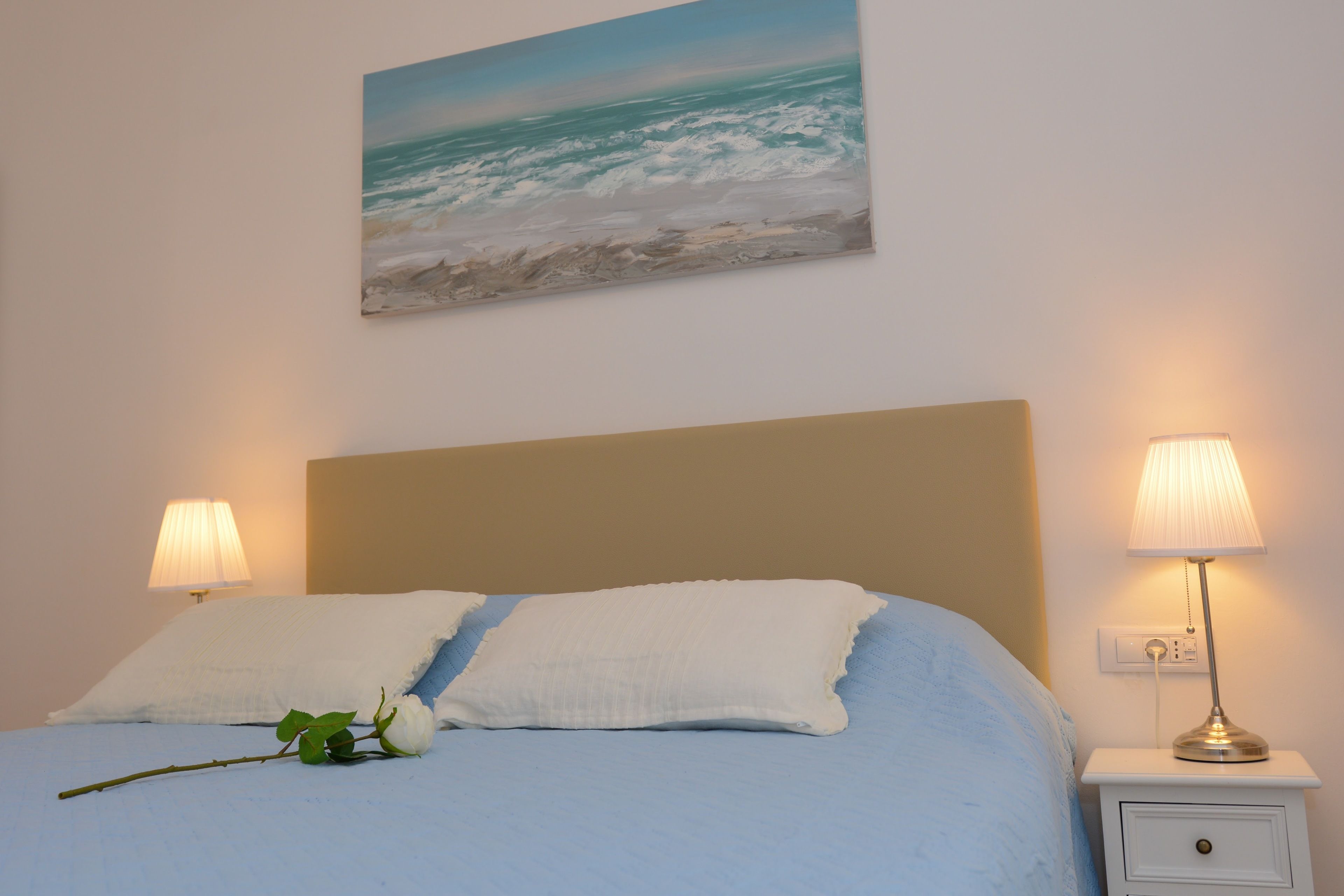 A neatly made bed with white linens, a single white pillow, and a rose on the bed. There is a painting of a beach scene above the bed. Two bedside lamps are on, and a small white nightstand with a lamp is to the right of the bed.