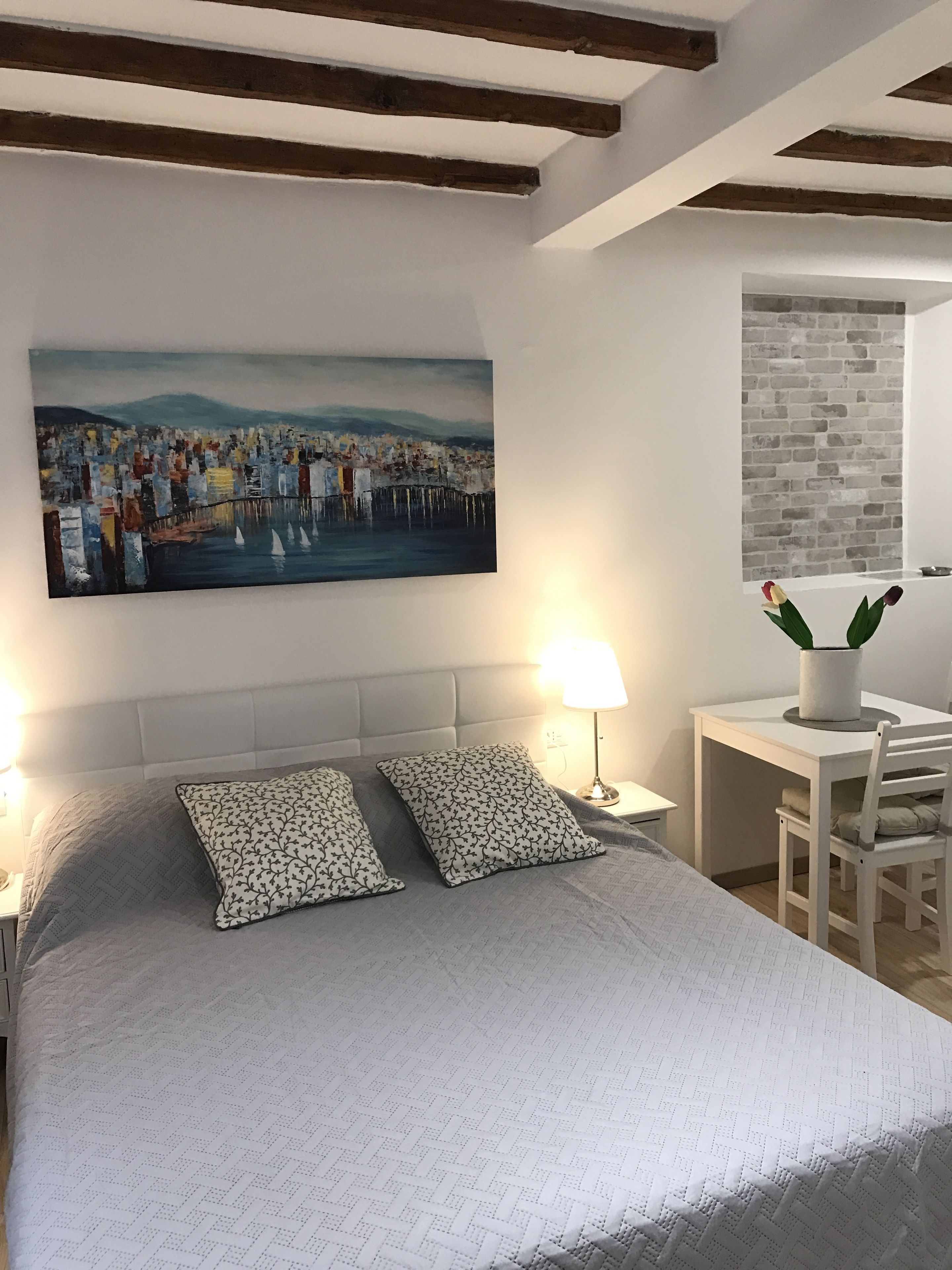 A bedroom with a large bed, two bedside lamps, a desk with a chair, and a painting on the wall.