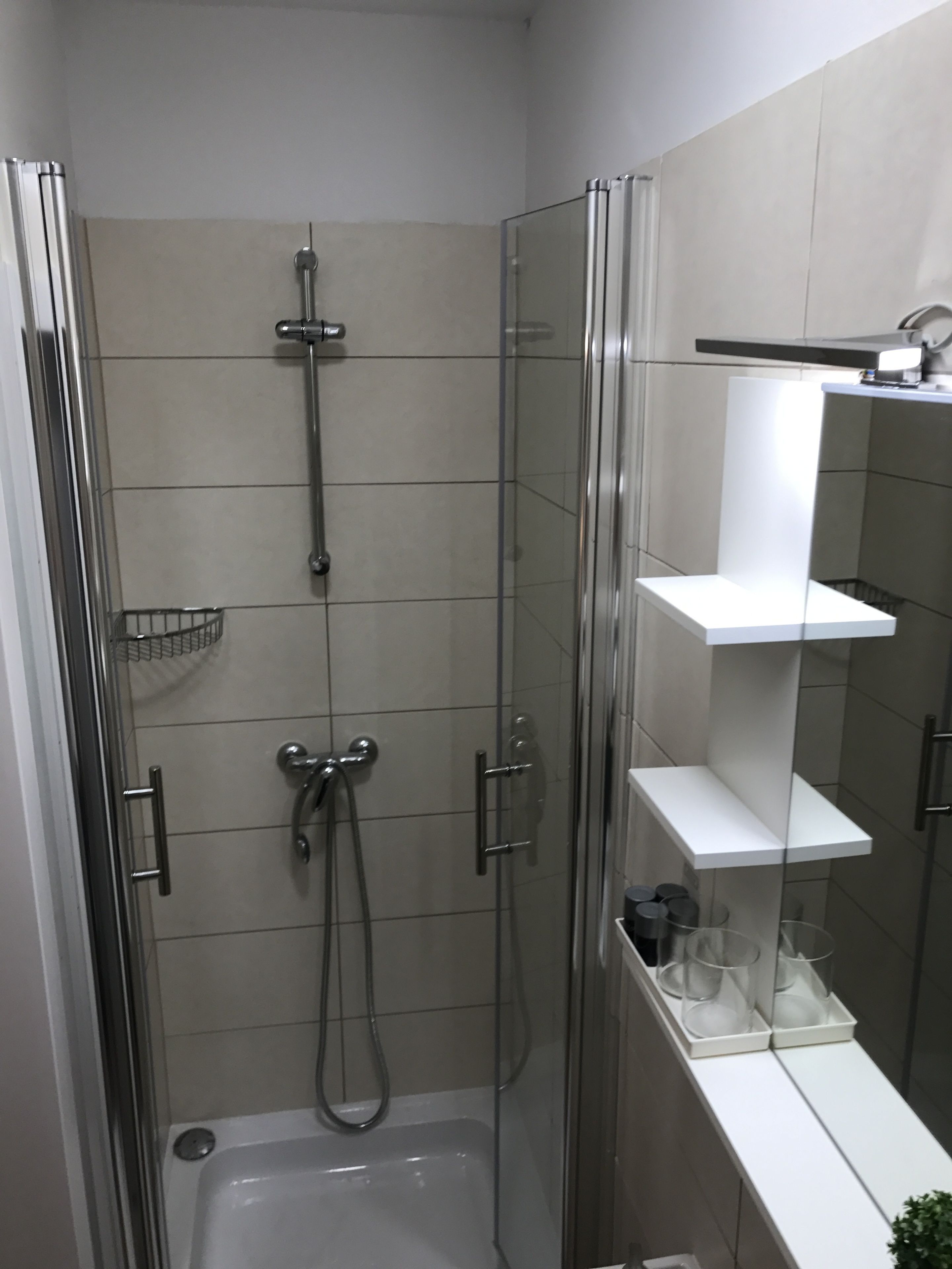 A glass-enclosed shower stall with a handheld showerhead, a wall-mounted faucet, and a small shelf with toiletries.