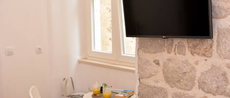 A room with a stone wall, a mounted TV, a small round table with a book, glasses, and a kettle.