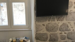 A room with a stone wall, a flat-screen TV mounted above a small table with glasses and a bag of cereal. There are two cushioned chairs and a bed with a quilted cover. - Rilati Old Town Palace (Dubrovnik)
