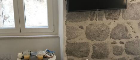 A room with a stone wall, a flat-screen TV mounted above a small table with glasses and a bag of cereal. There are two cushioned chairs and a bed with a quilted cover.