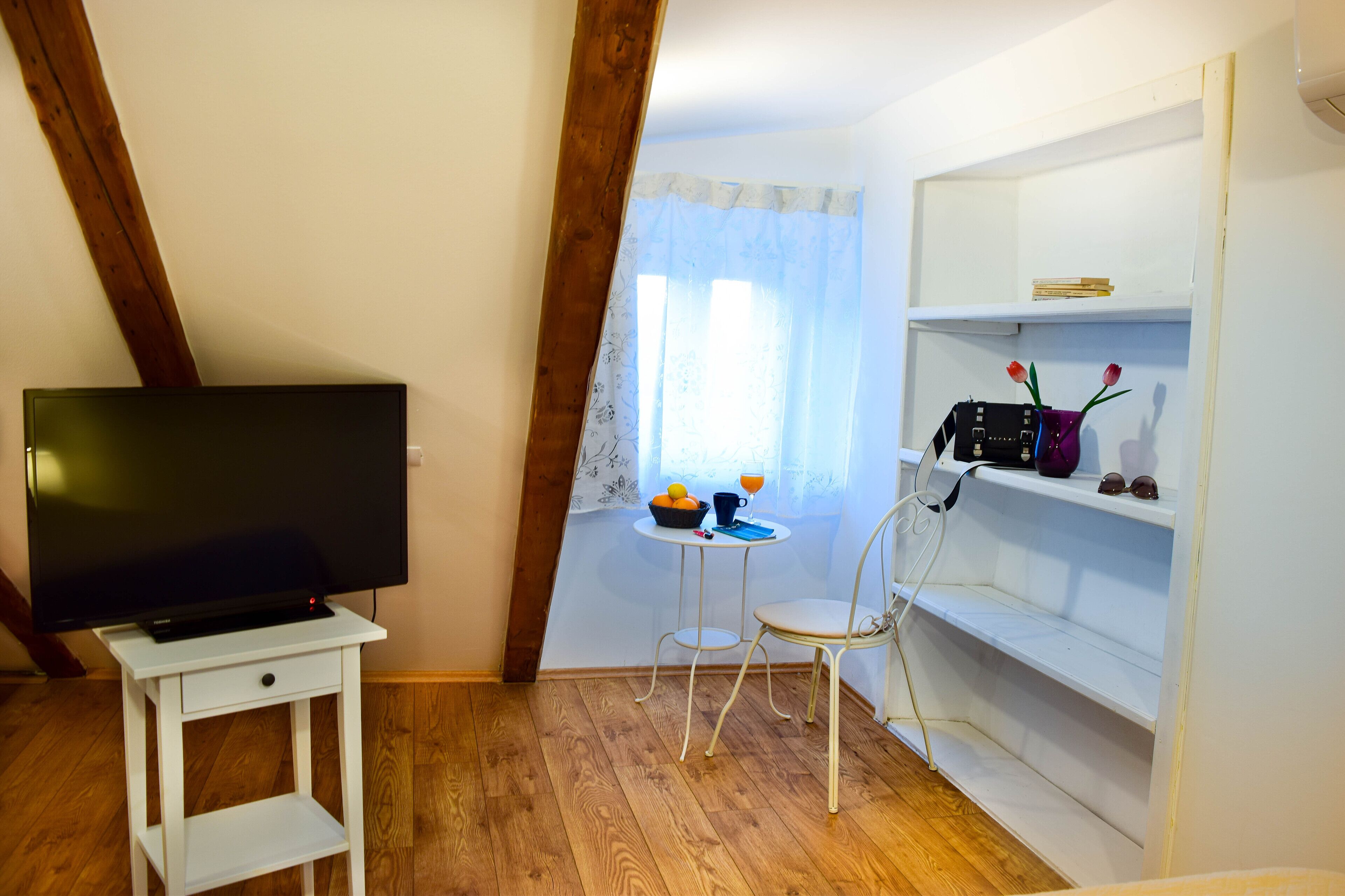 Una habitación con una televisión sobre un mueble blanco, una mesita con frutas y una taza, una silla blanca y un estante blanco con una radio y elementos decorativos.