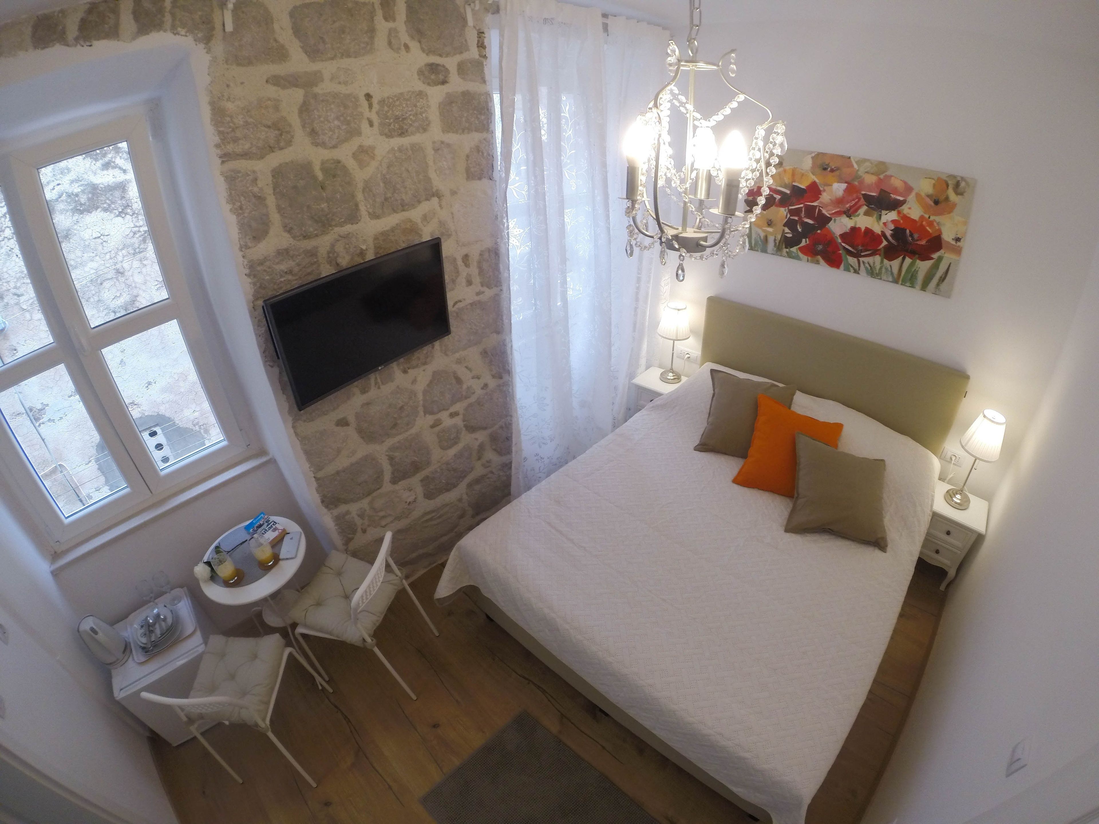 A bedroom with a stone wall, a flat-screen TV, a bed with white and orange pillows, and a chandelier.