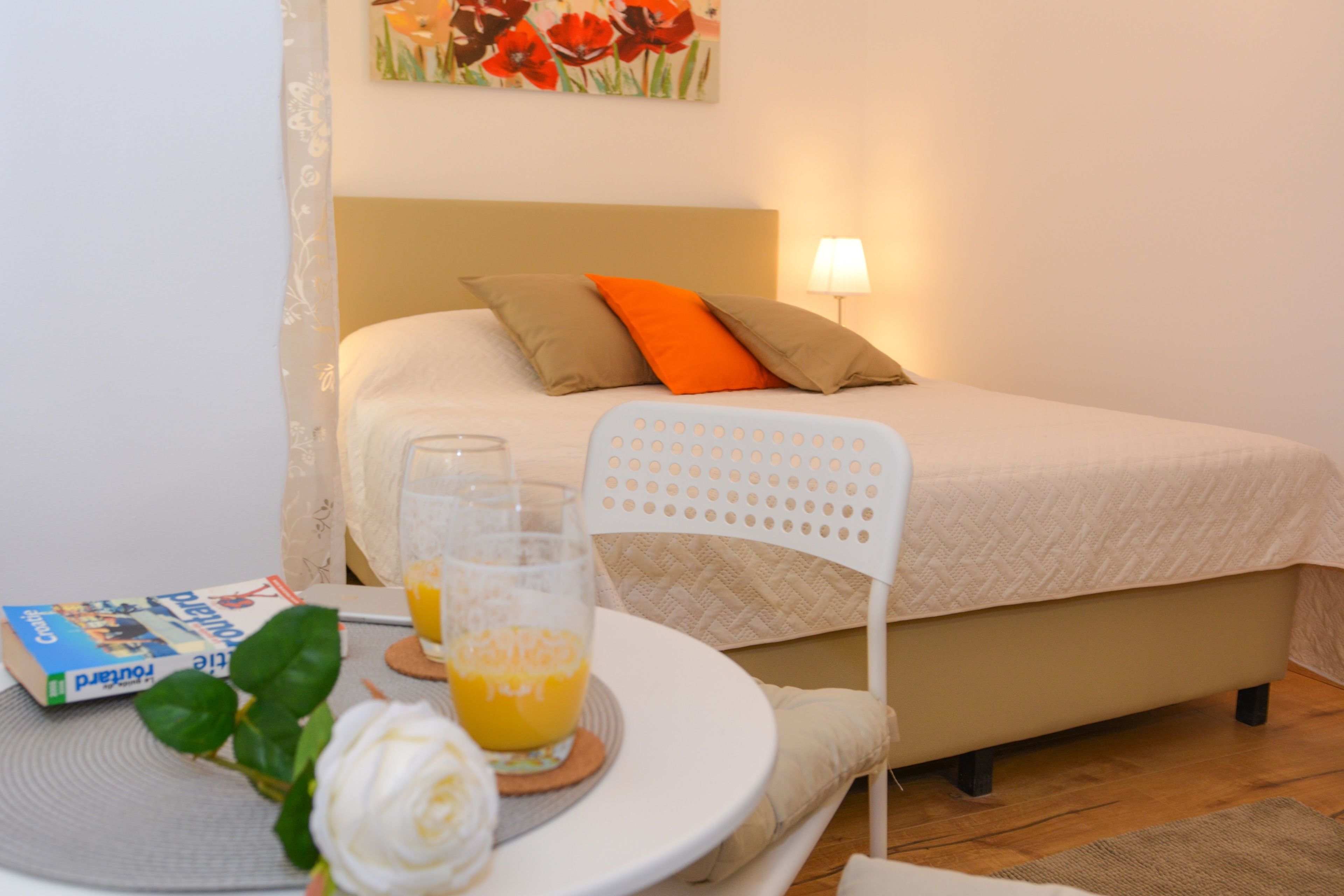 Quarto bem arrumado com uma cama, travesseiros, uma mesa de cabeceira com um copo de suco de laranja, um livro e uma cadeira branca.