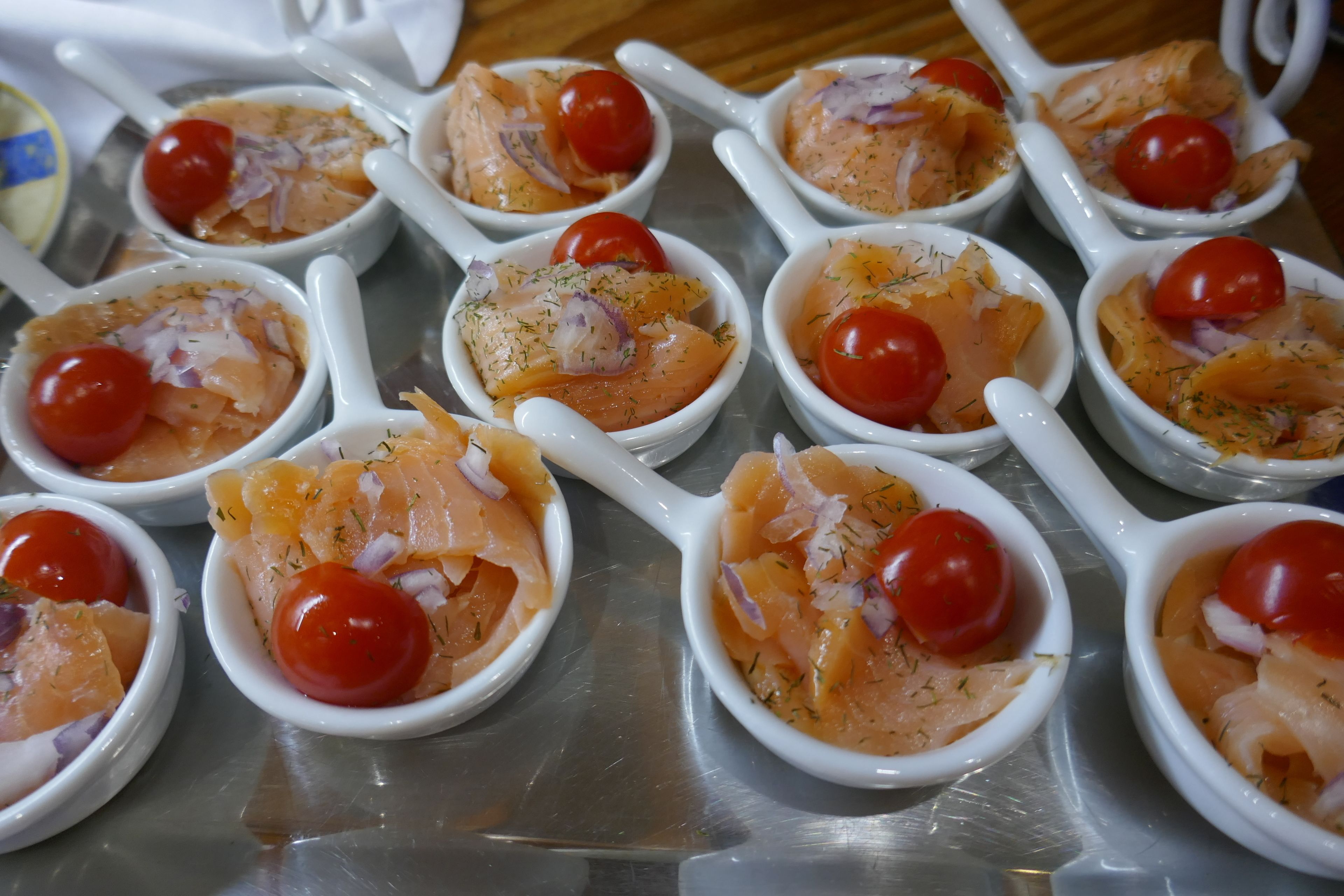 Mehrere weiße Schalen mit Lachs und Kirschtomaten auf einer Metalltablett.