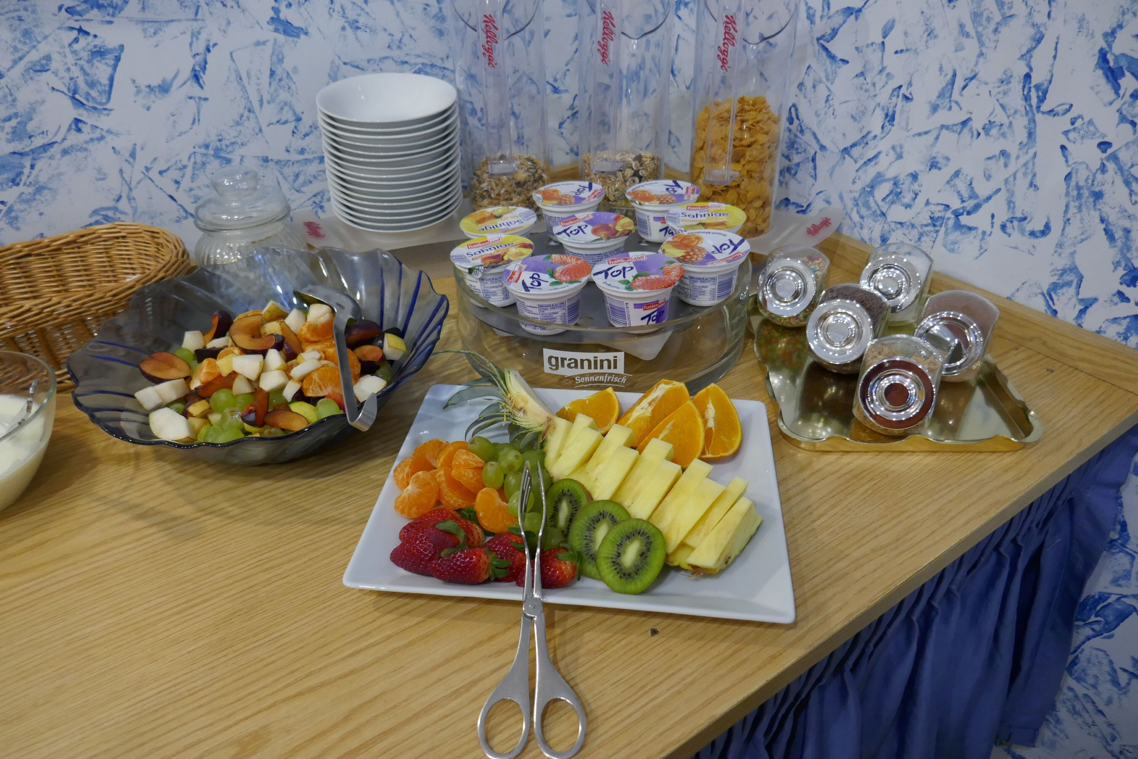 Una mesa de buffet con diversas opciones de comida, incluyendo frutas, Granini y vasos de yogur.
