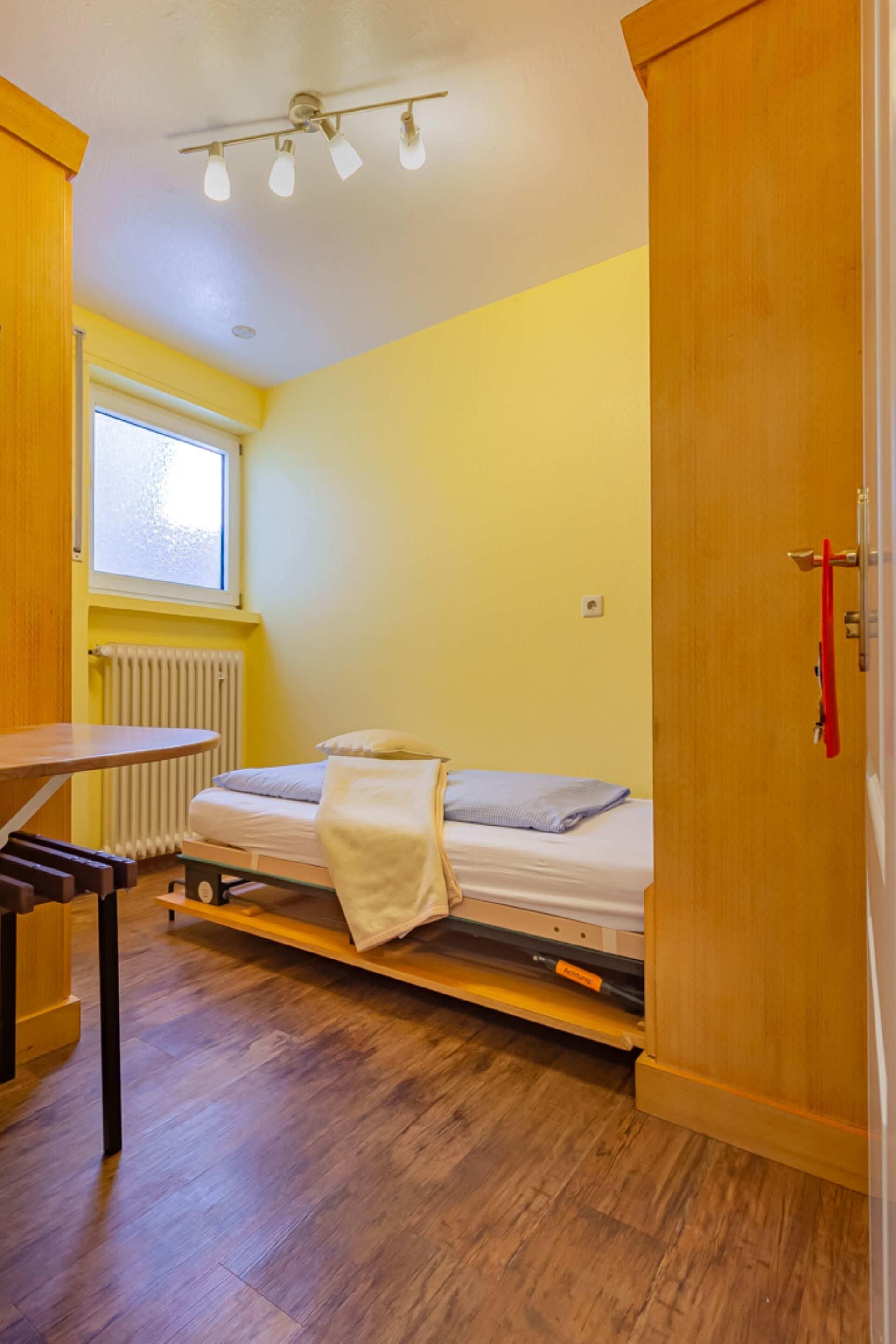 A small, bright room with a bed, a table, and a radiator.