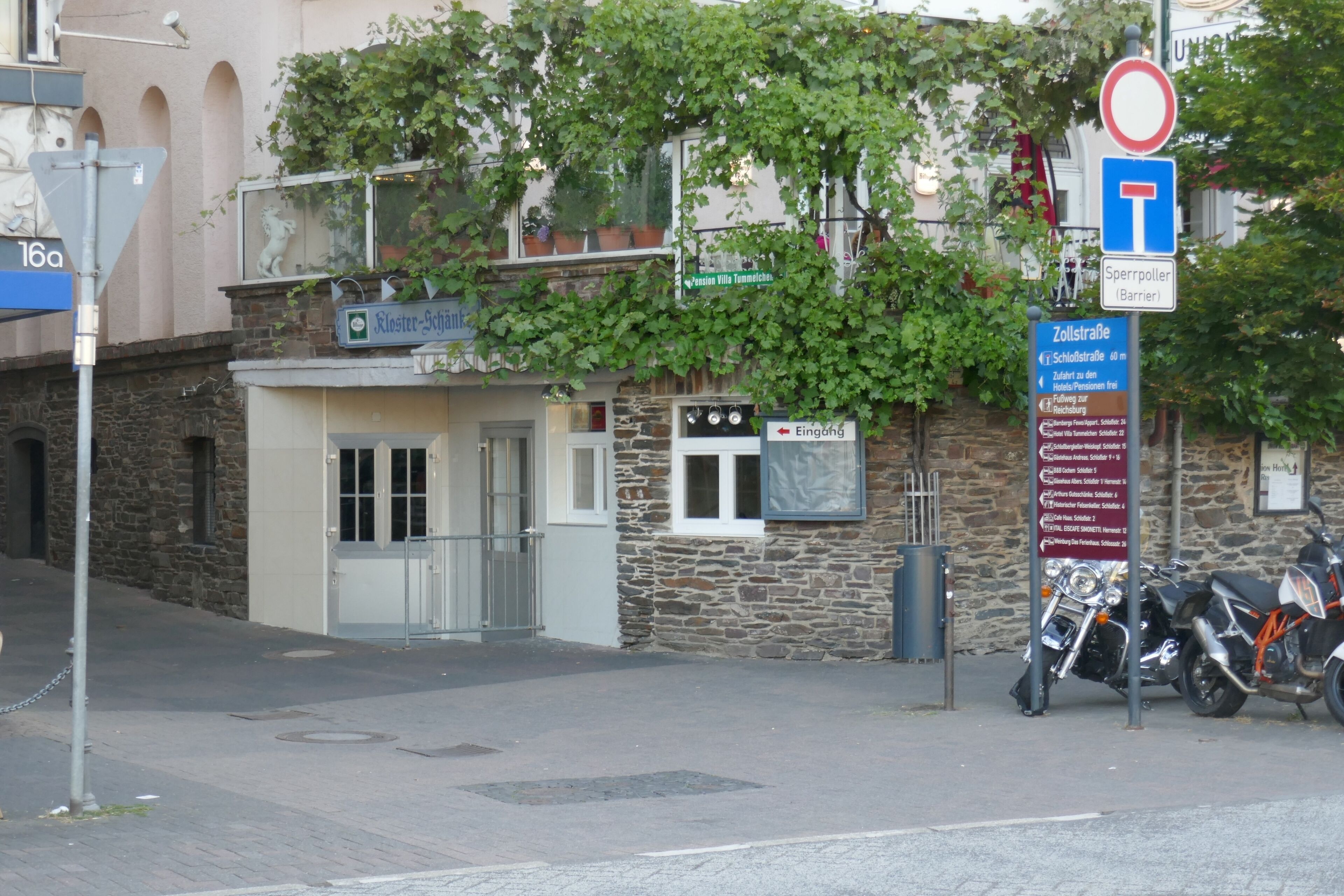 A building with a stone facade, a sign indicating 'Eingang' (Entrance), and a motorcycle parked outside.