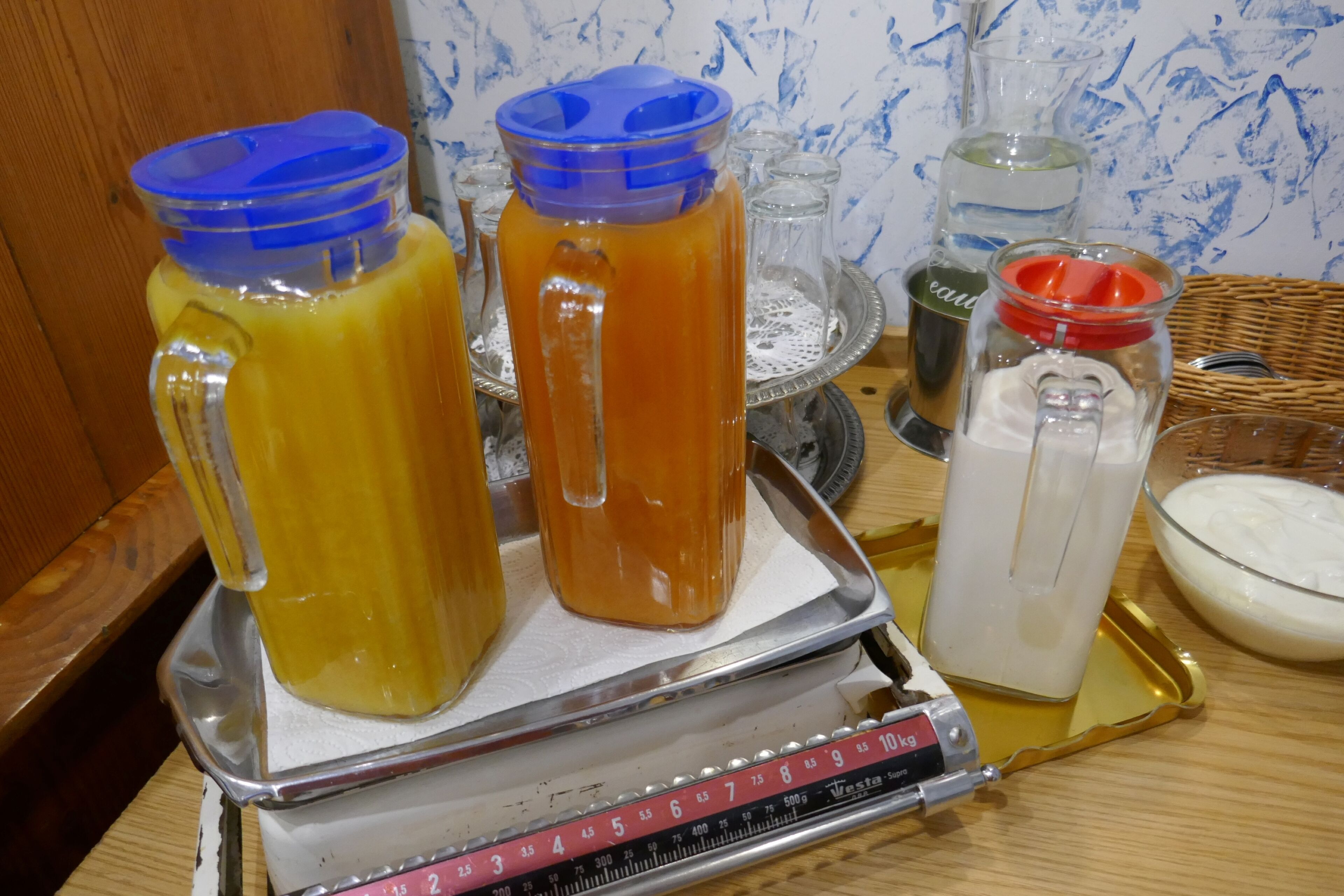 A kitchen counter with two large glass jars with blue lids, a metal scale, a glass of milk, and a bowl of cream.