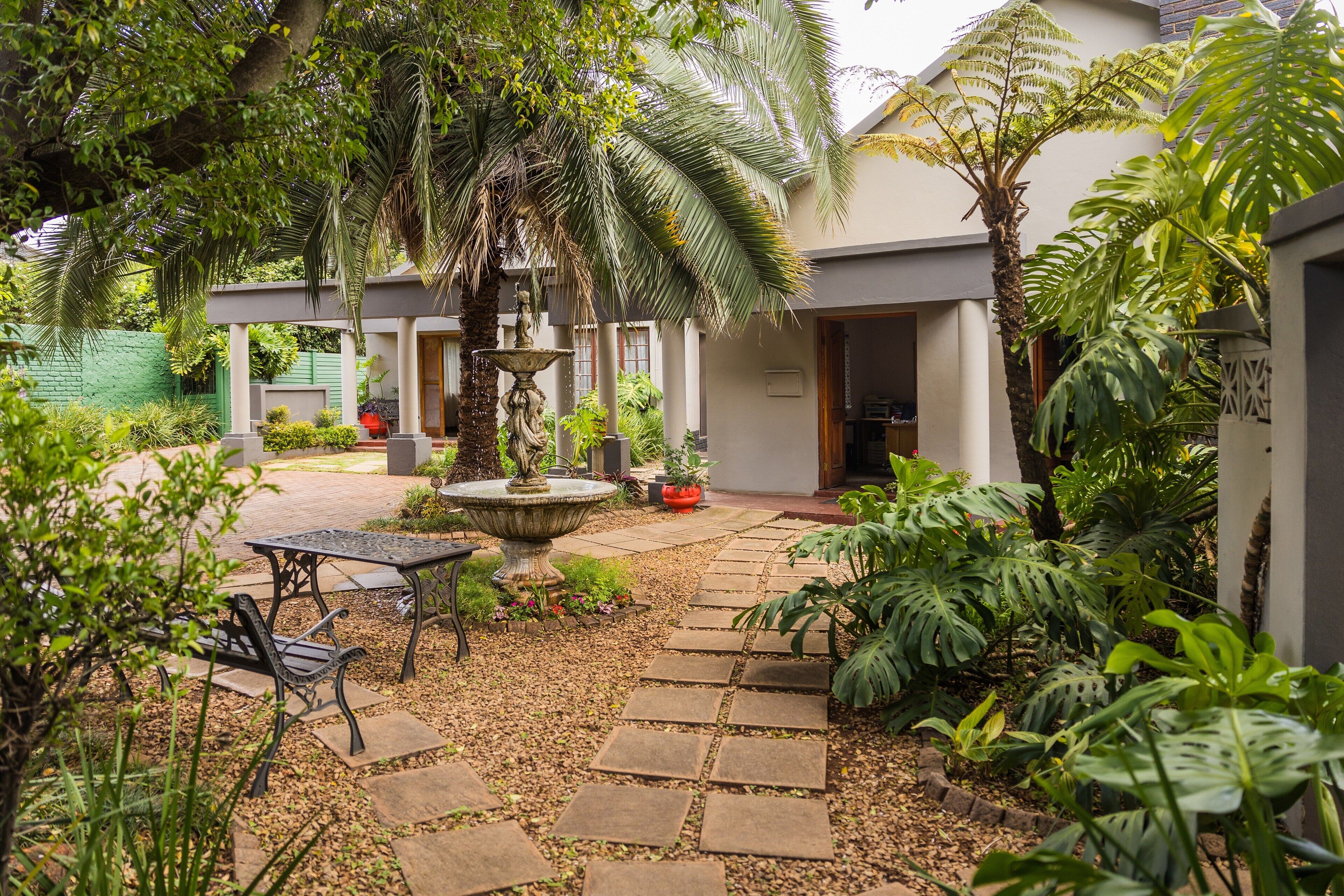 A courtyard with a stone pathway, a table with chairs, a fountain, and various tropical plants.