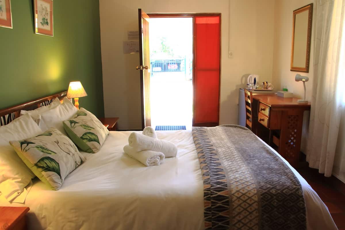 A neatly made bed with white linens and green leaf-patterned pillows, a bedside lamp, a wooden dresser with a mirror, and a red door leading outside.