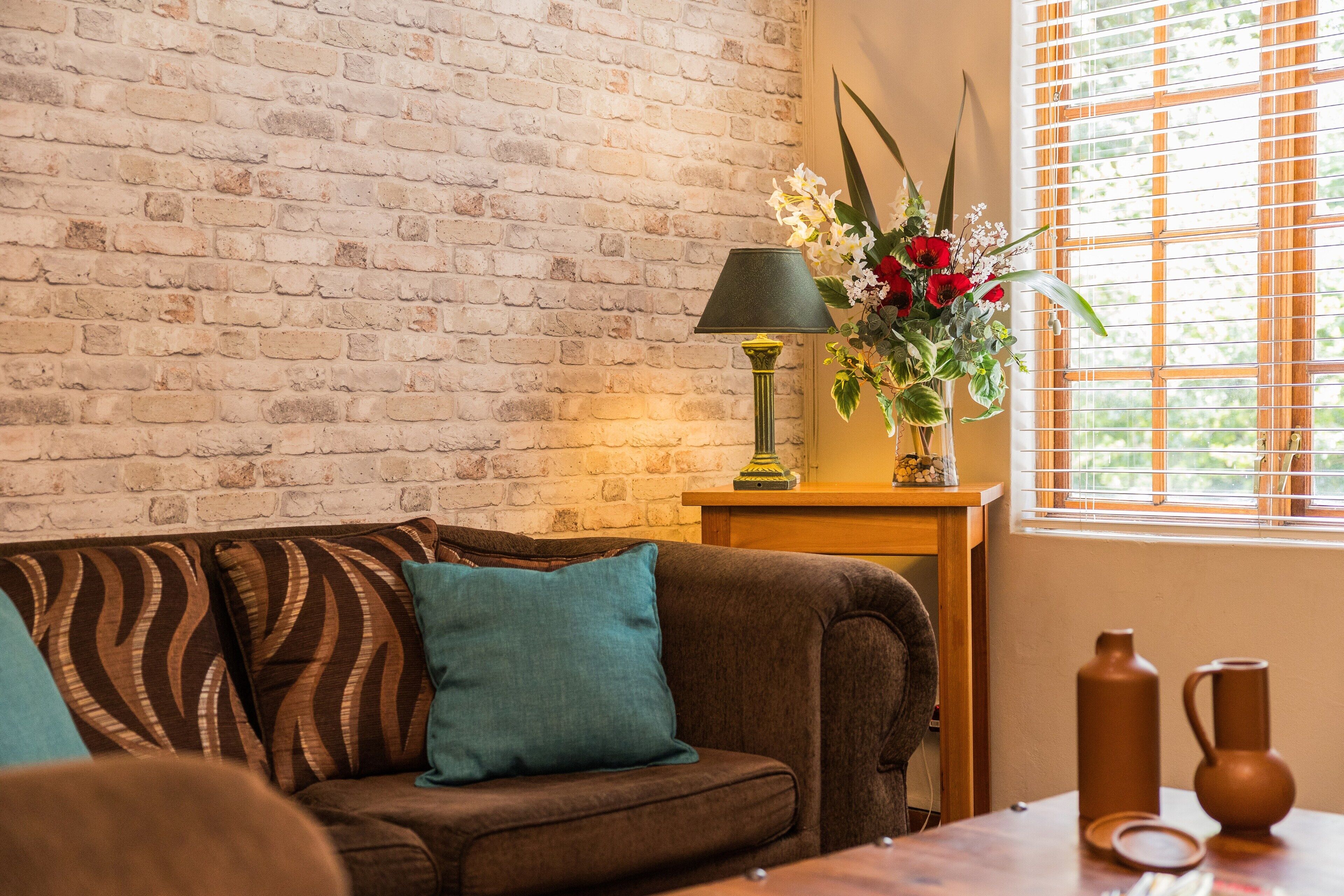 A cozy living room with a brick wall, a wooden side table with a lamp, a vase of flowers, and a window with blinds.