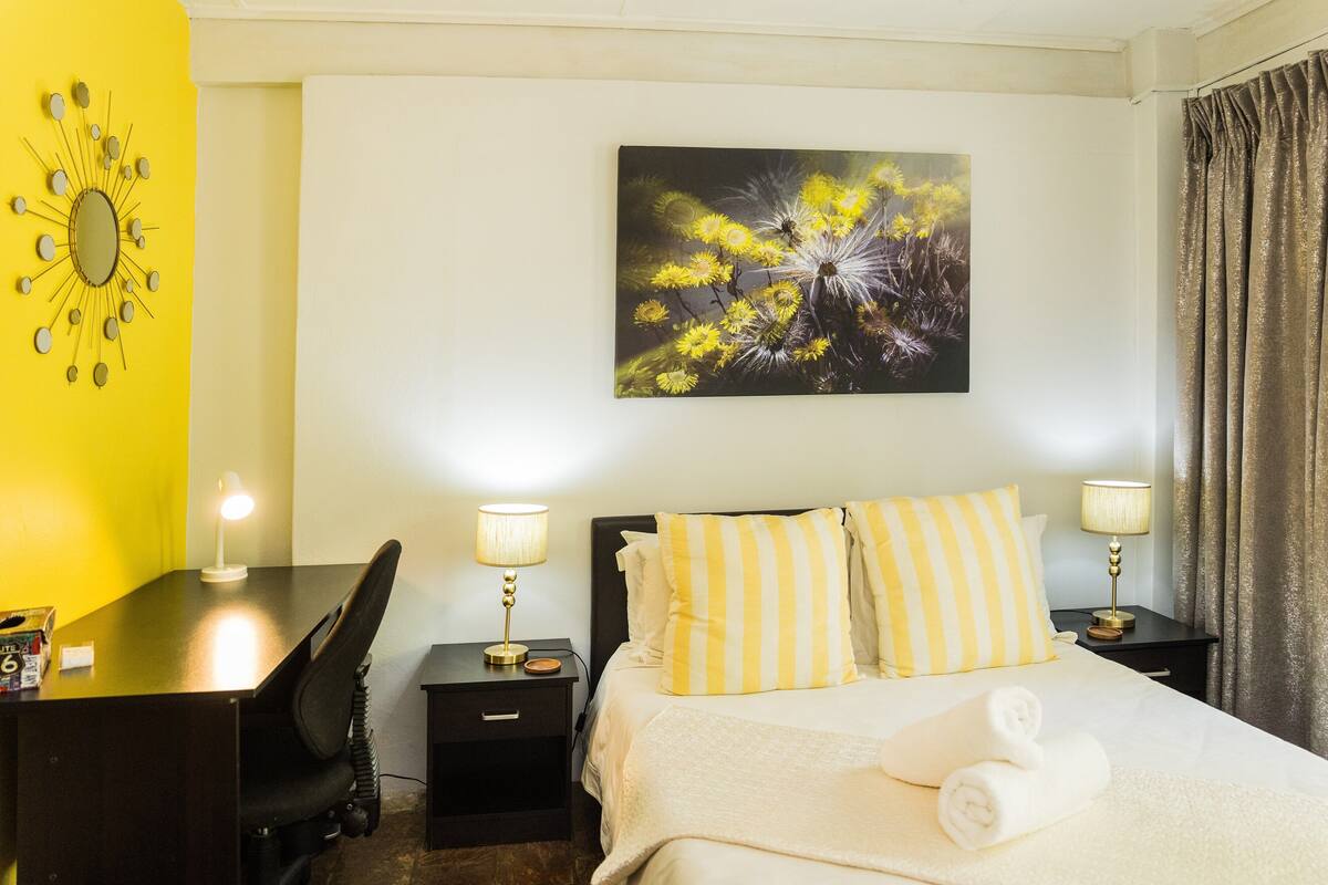 A hotel room with a bed, two bedside tables with lamps, a desk, a chair, a wall clock, and a painting of flowers.