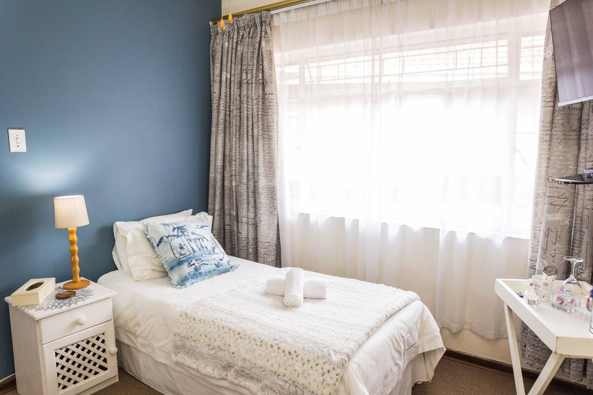 A bedroom with a bed, a bedside table with a lamp, a small desk with toiletries, and curtains.