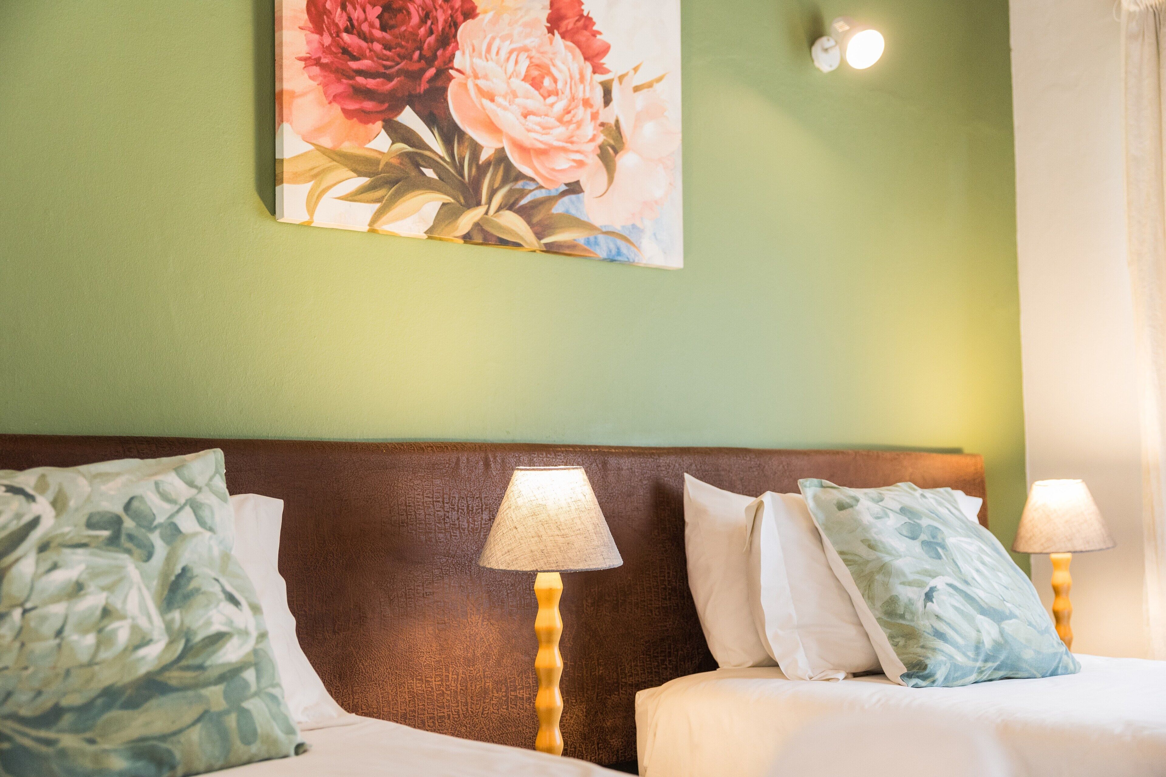 Habitación de hotel con pared verde, un cuadro floral, dos lámparas de noche y una cama con ropa blanca y almohadas estampadas.