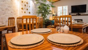 Un comedor con sillas de madera y mesas redondas, pared de ladrillo y una planta grande.