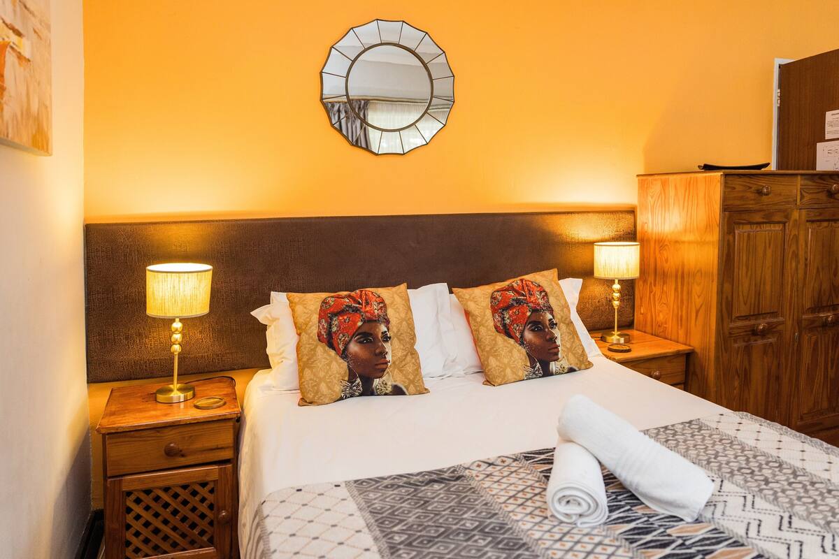 A hotel room with a bed, two bedside lamps, a wooden headboard, a mirror, a wooden wardrobe, and a patterned bedspread.