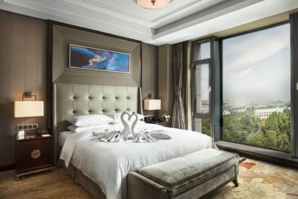 A hotel room with a large bed, a view of the city, a painting on the wall, and a nightstand with a lamp.