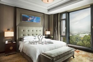A hotel room with a large bed, a view of the city, a painting on the wall, and a nightstand with a lamp.