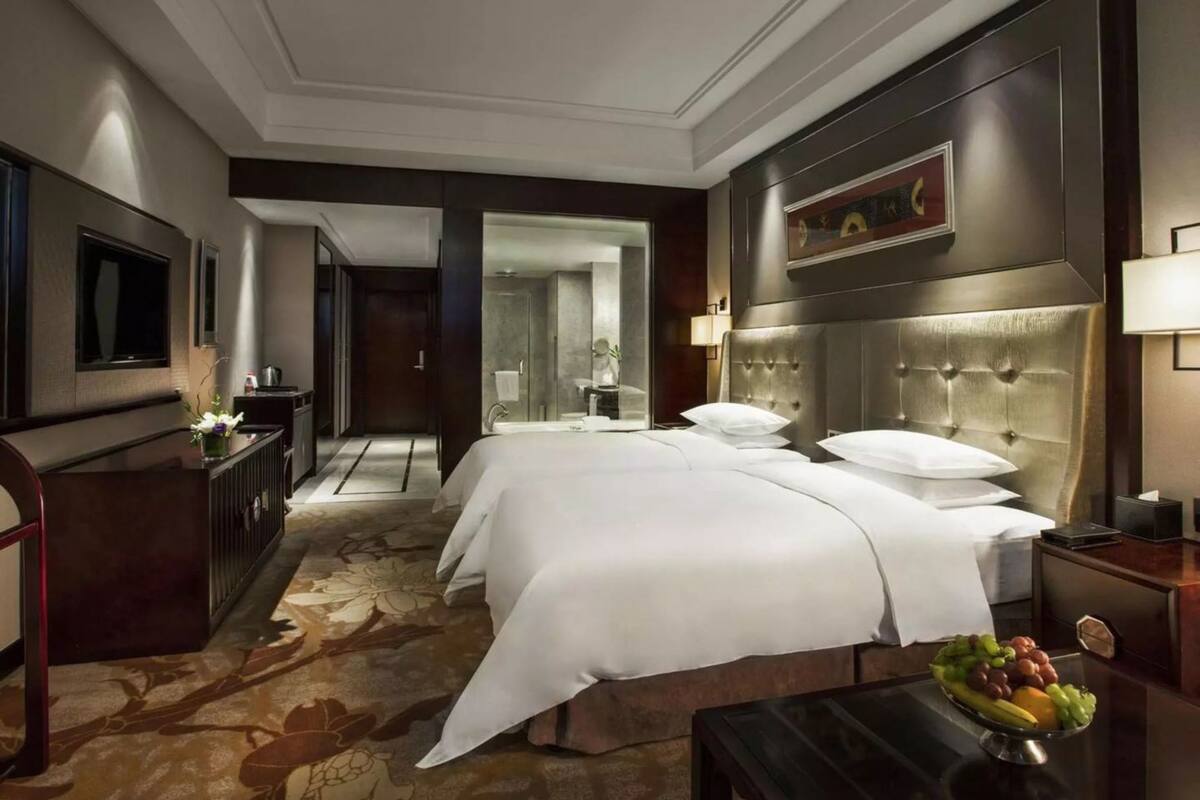 A hotel room with two beds, a large mirror, a flat-screen TV, a fruit bowl, and a decorative wall piece.