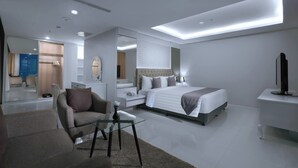 Moderni hotellihuone, jossa on suuri sänky, oleskelualue lasipöydällä, taulutelevisio ja suuri peili.