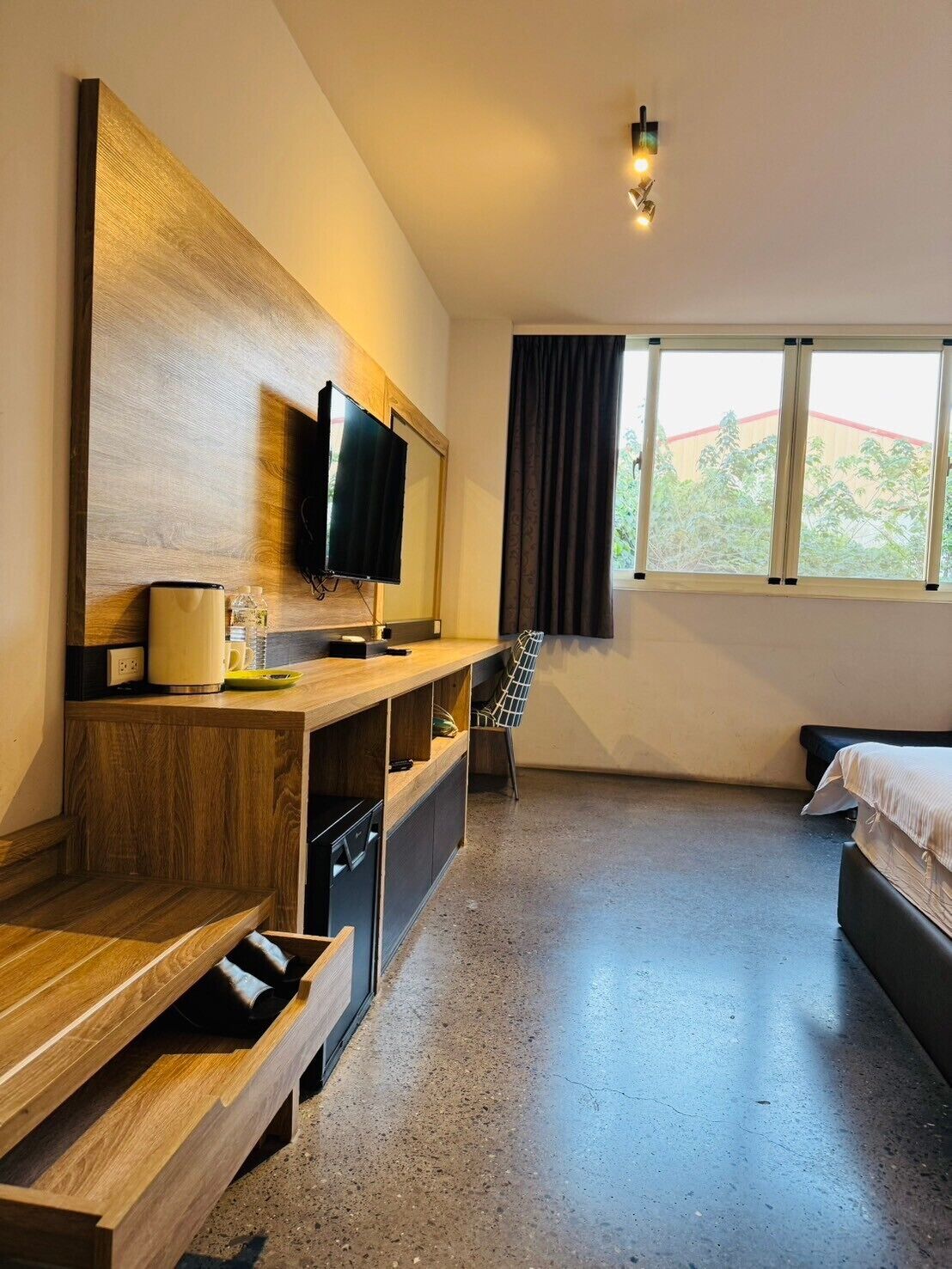 A hotel room with a wooden TV stand, a flat-screen TV, a bed, a chair, and a window with curtains.