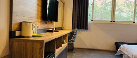 A hotel room with a wooden TV stand, a flat-screen TV, a bed, a chair, and a window with curtains.