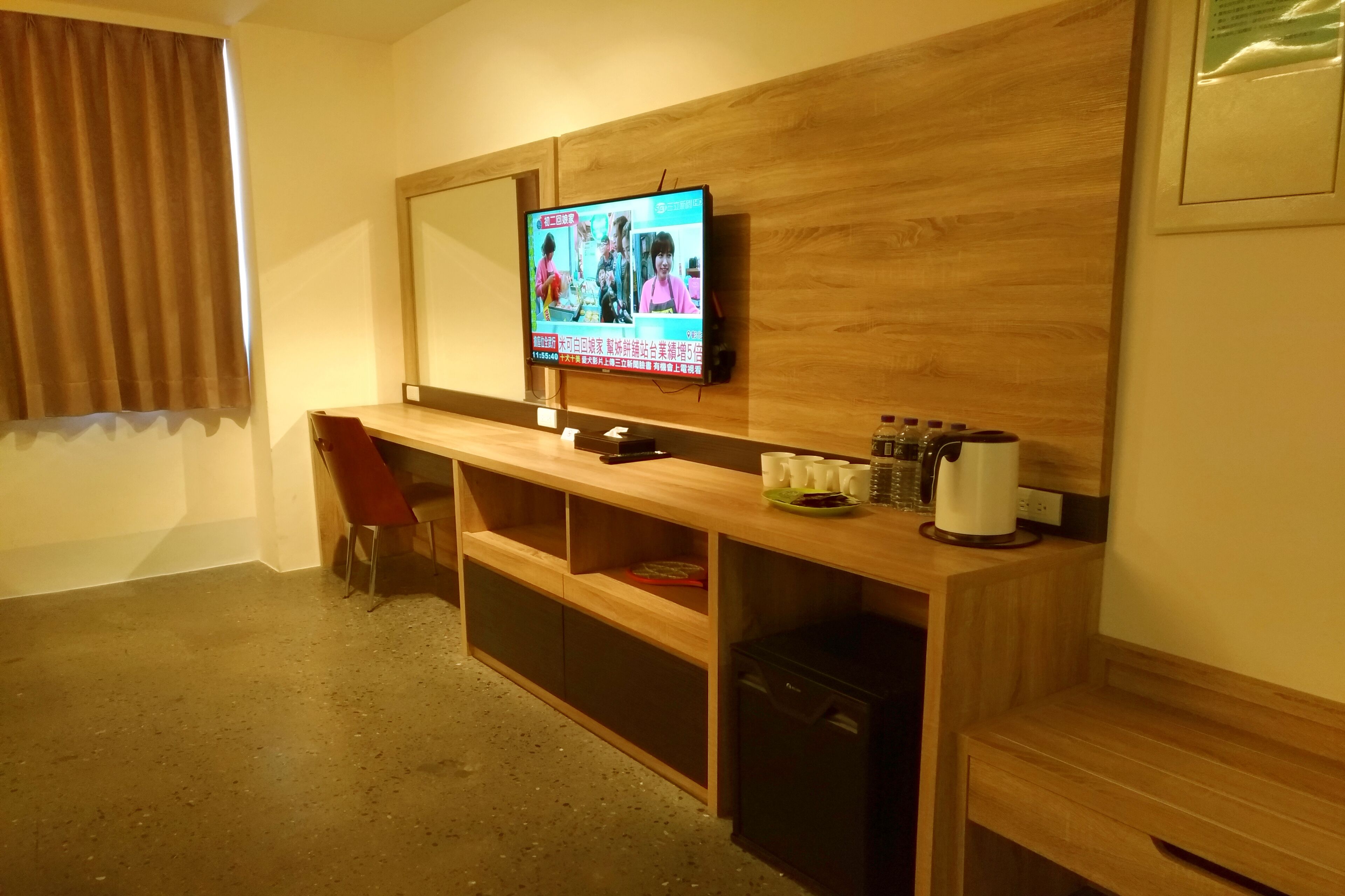 A hotel room with a wooden desk, a television mounted on the wall, a red chair, and a small cabinet.