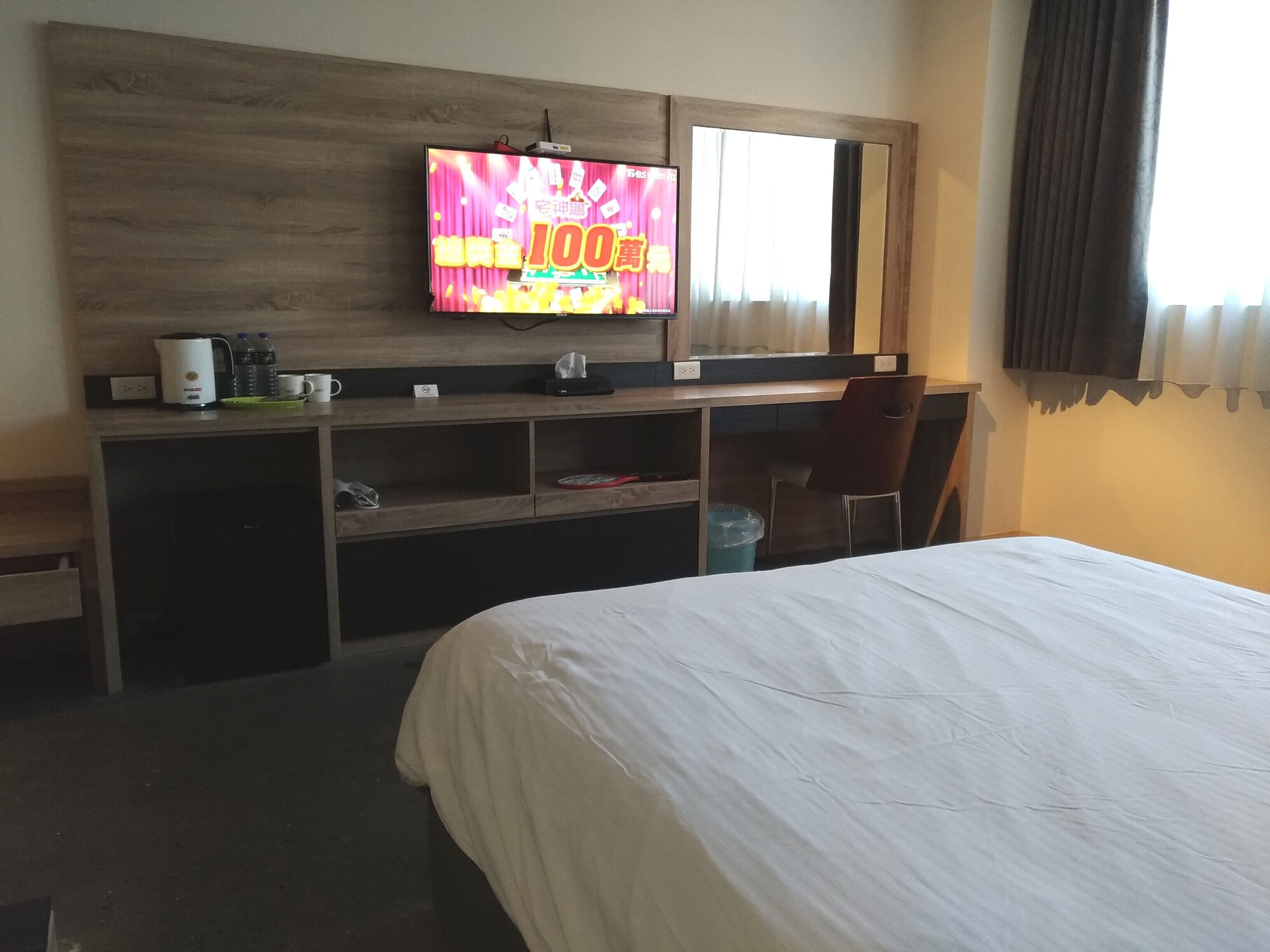 A hotel room with a bed, a TV mounted on the wall, a desk with a chair, and a window with curtains.