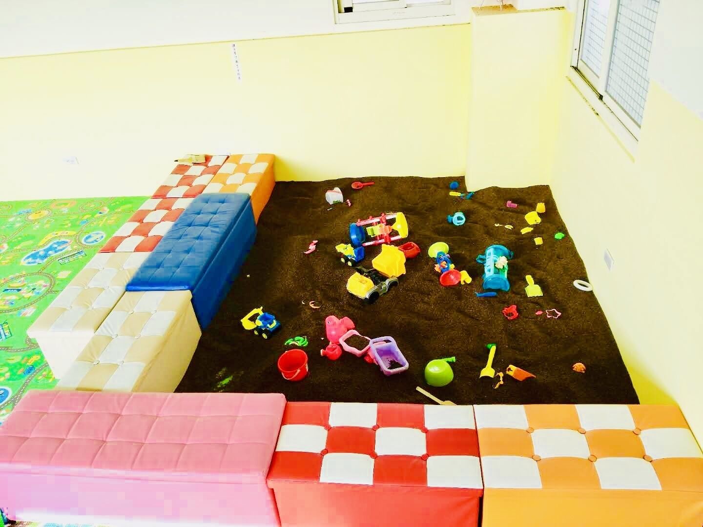 Uma área de recreação com estrutura de brinquedos macios, tapete colorido e diversos brinquedos espalhados pelo chão.