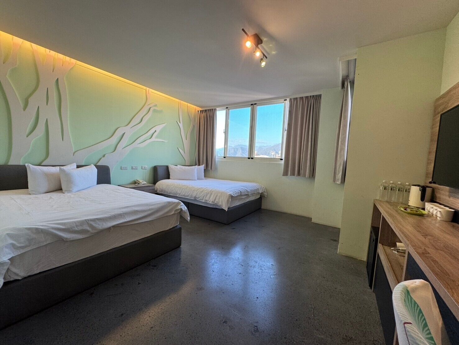 Habitación de hotel con dos camas, un escritorio con silla, un ventanal con cortinas y una pared con diseño arbóreo.