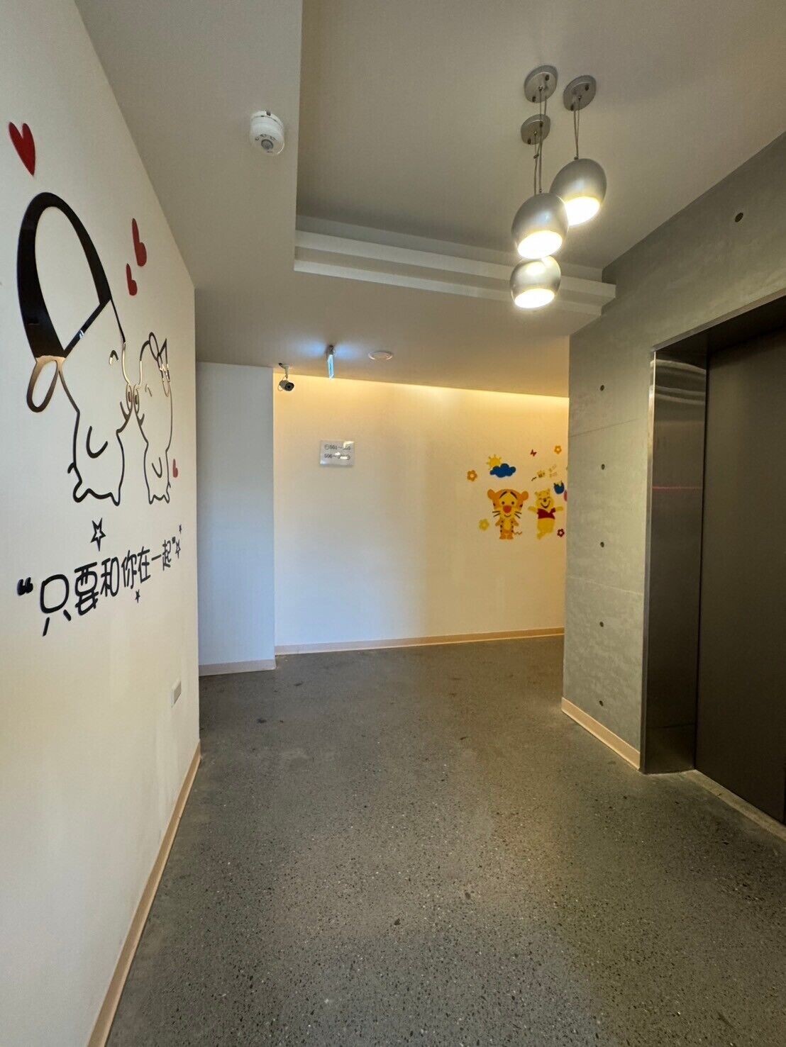 A hallway with a concrete floor, a wall with a cartoon character and text, a ceiling with three pendant lights, and an elevator.