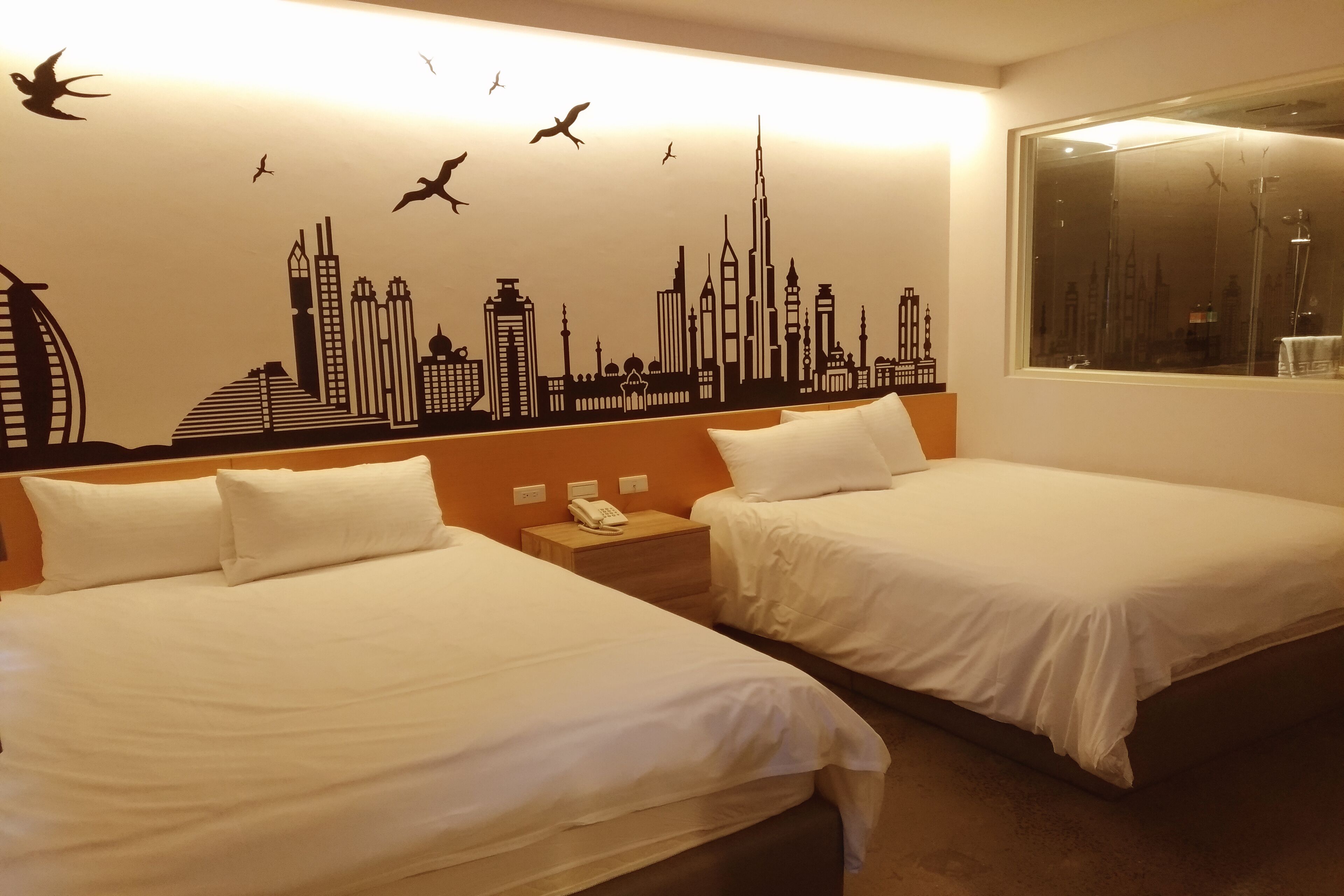 A hotel room with two beds, a cityscape wall mural, a window with a view, and a nightstand with a phone.