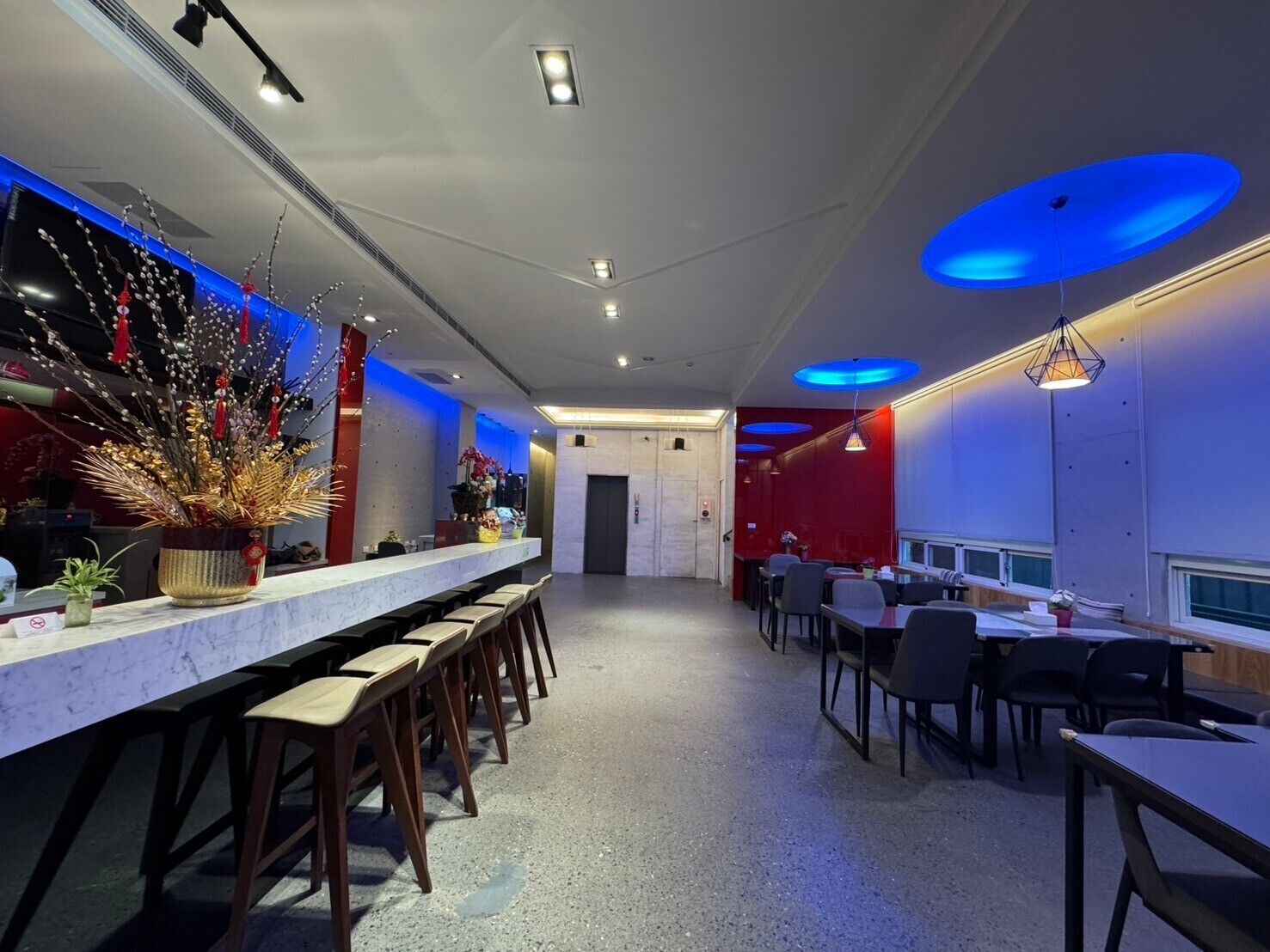 En moderne bar med en lang bardisk, flere barstole og borde. Der er dekorative planter og en rød væg.