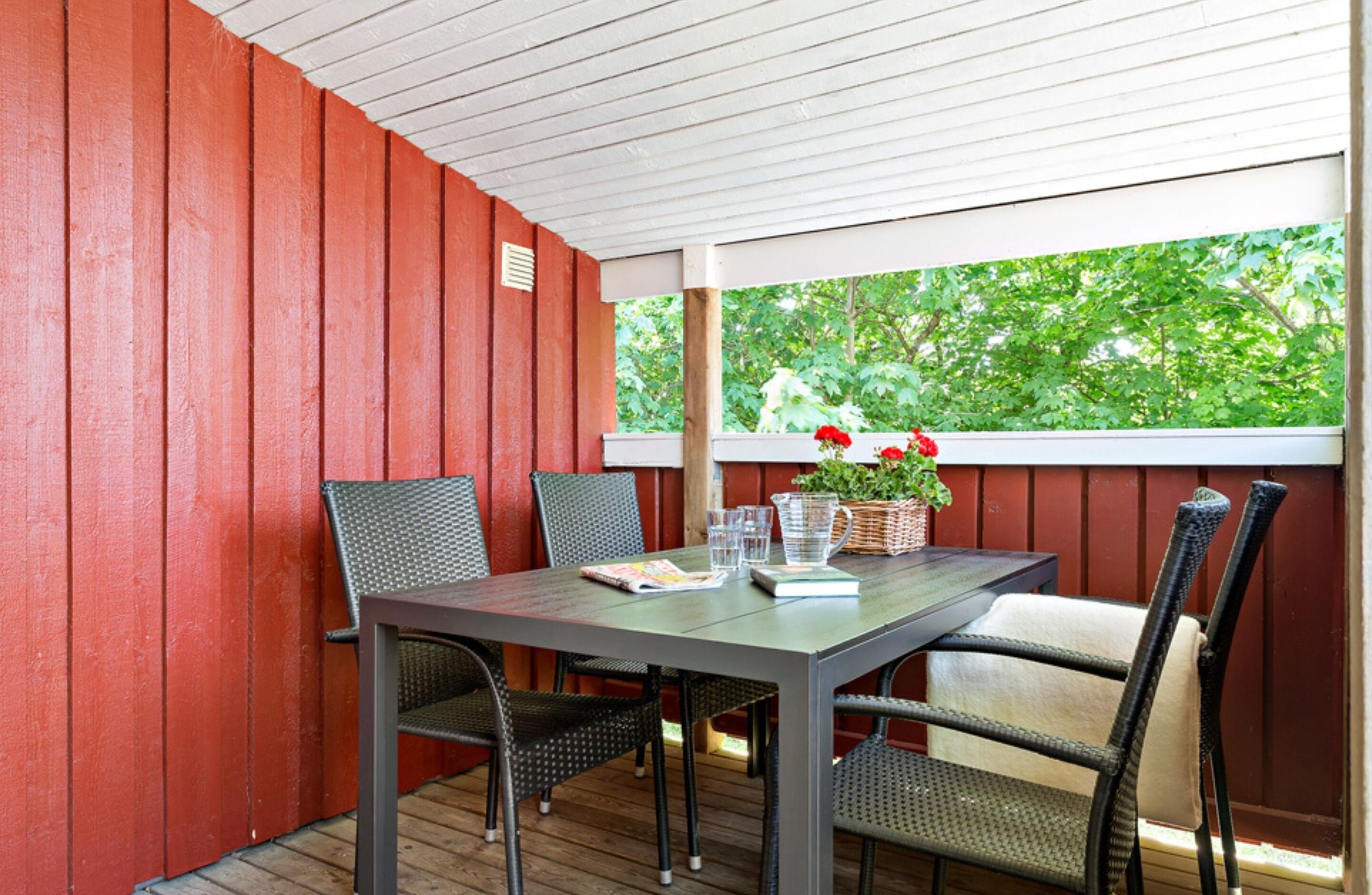Une terrasse couverte avec une table et des chaises, une plante en pot et une fenêtre donnant sur les arbres.