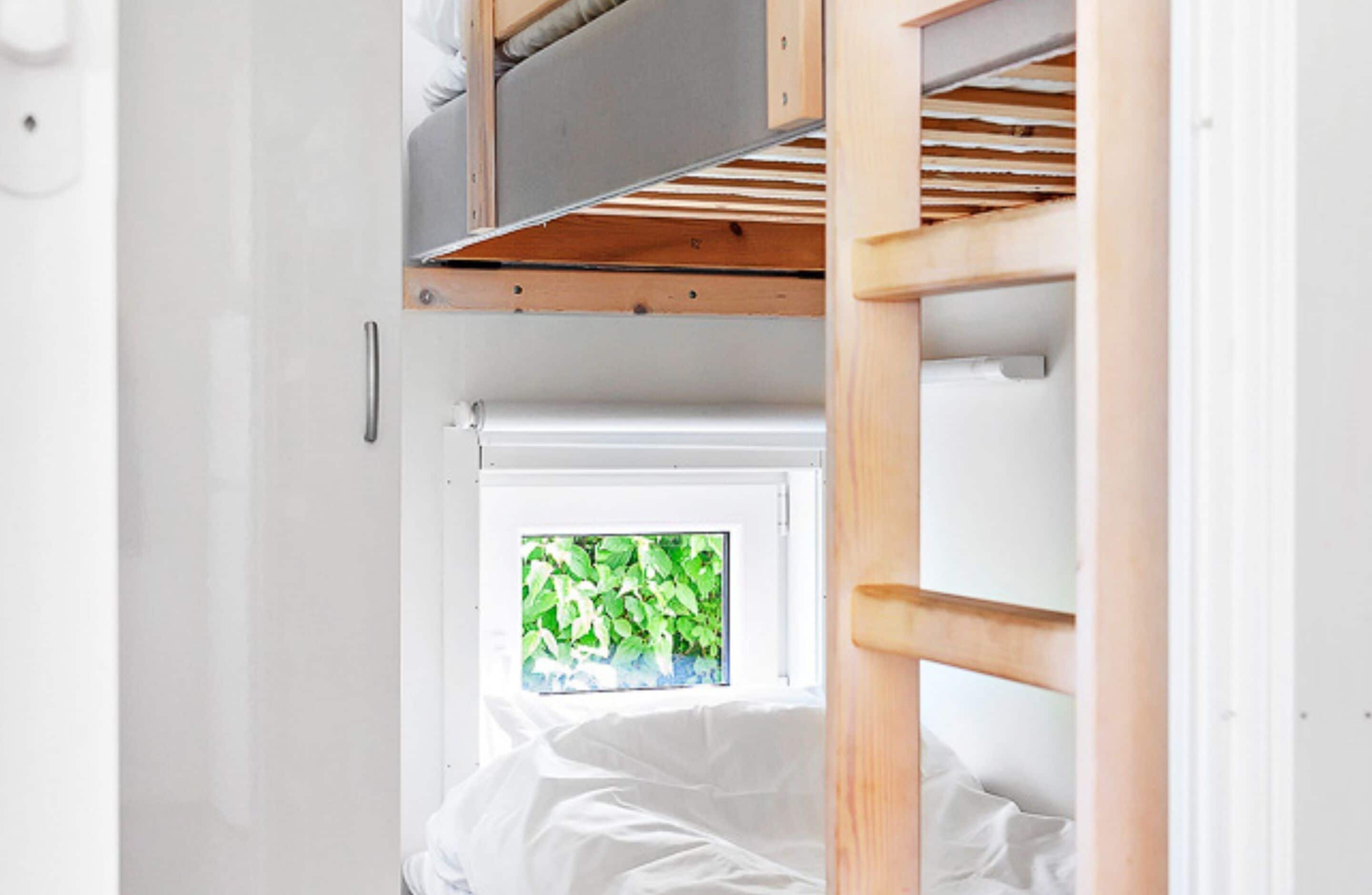 Een stapelbed met raam, witte muren en een houten trap.