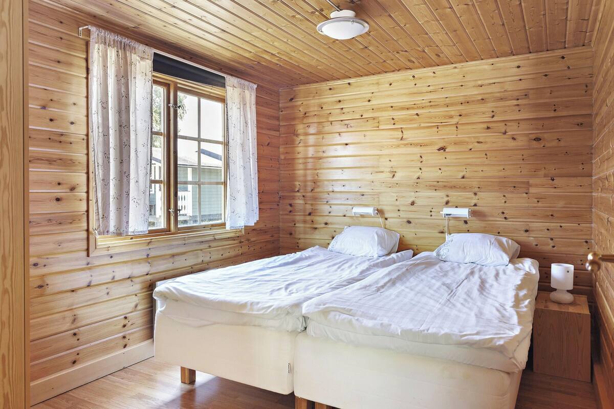 A wooden-paneled room with a bed, a window with curtains, and a bedside table with a lamp.