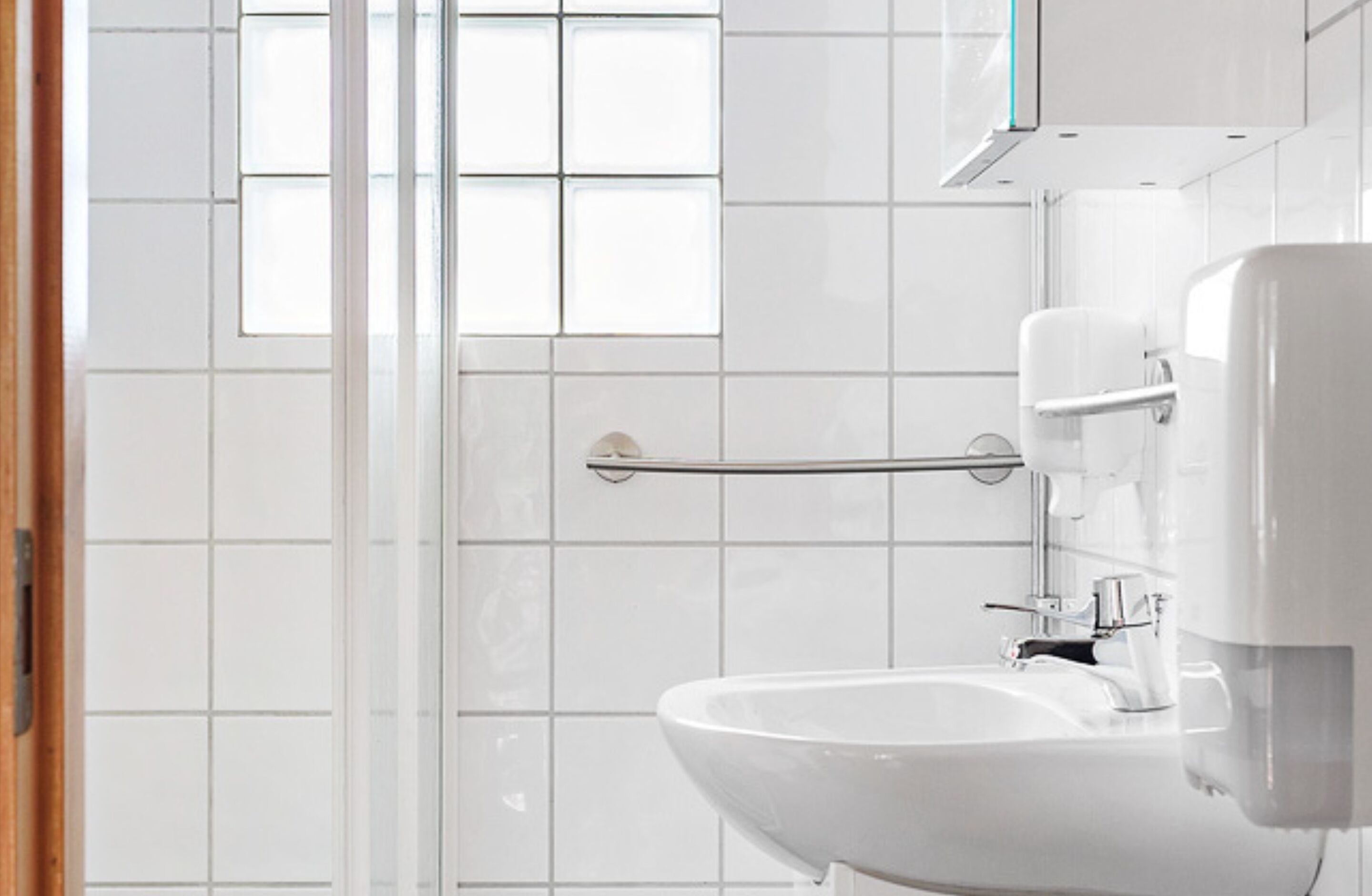 Une salle de bain avec des carreaux blancs, une porte de douche en verre, une barre d’appui, un lavabo et des toilettes.