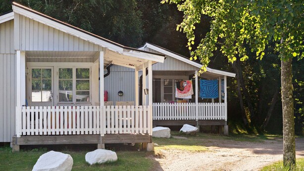 A cabin with a porch and a picket fence, surrounded by rocks and trees. - First Camp Tylösand (Halmstad)