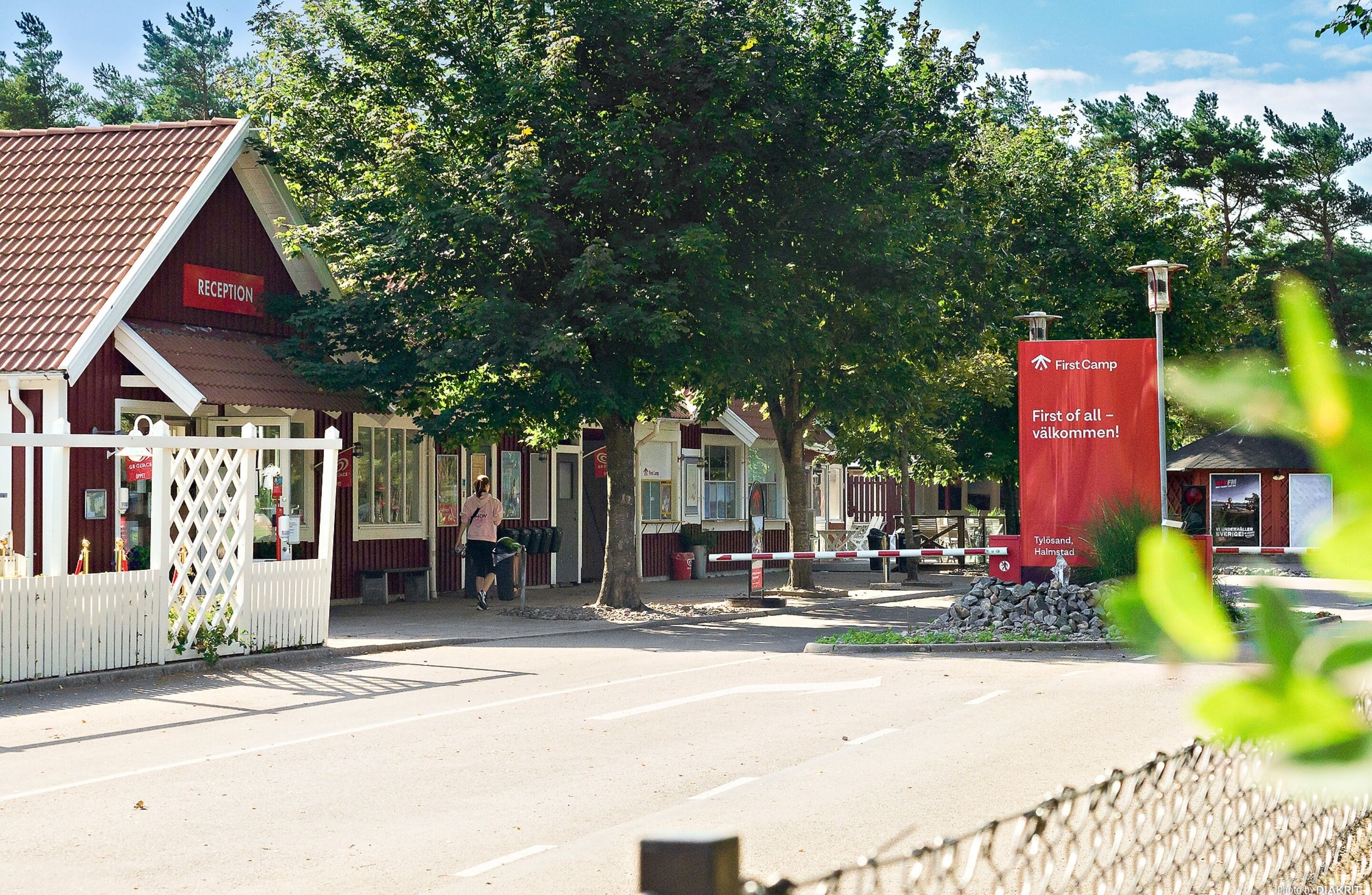 Ett receptionsområde med en röd byggnad, ett vitt staket och en skylt som visar att det är ”First Camp”.