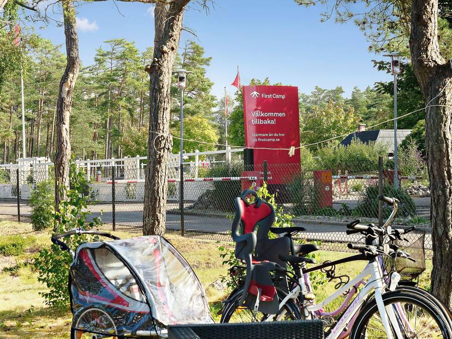 Um sinal vermelho com texto branco indicando direções para 'First Camp' e uma bicicleta estacionada perto de uma árvore.