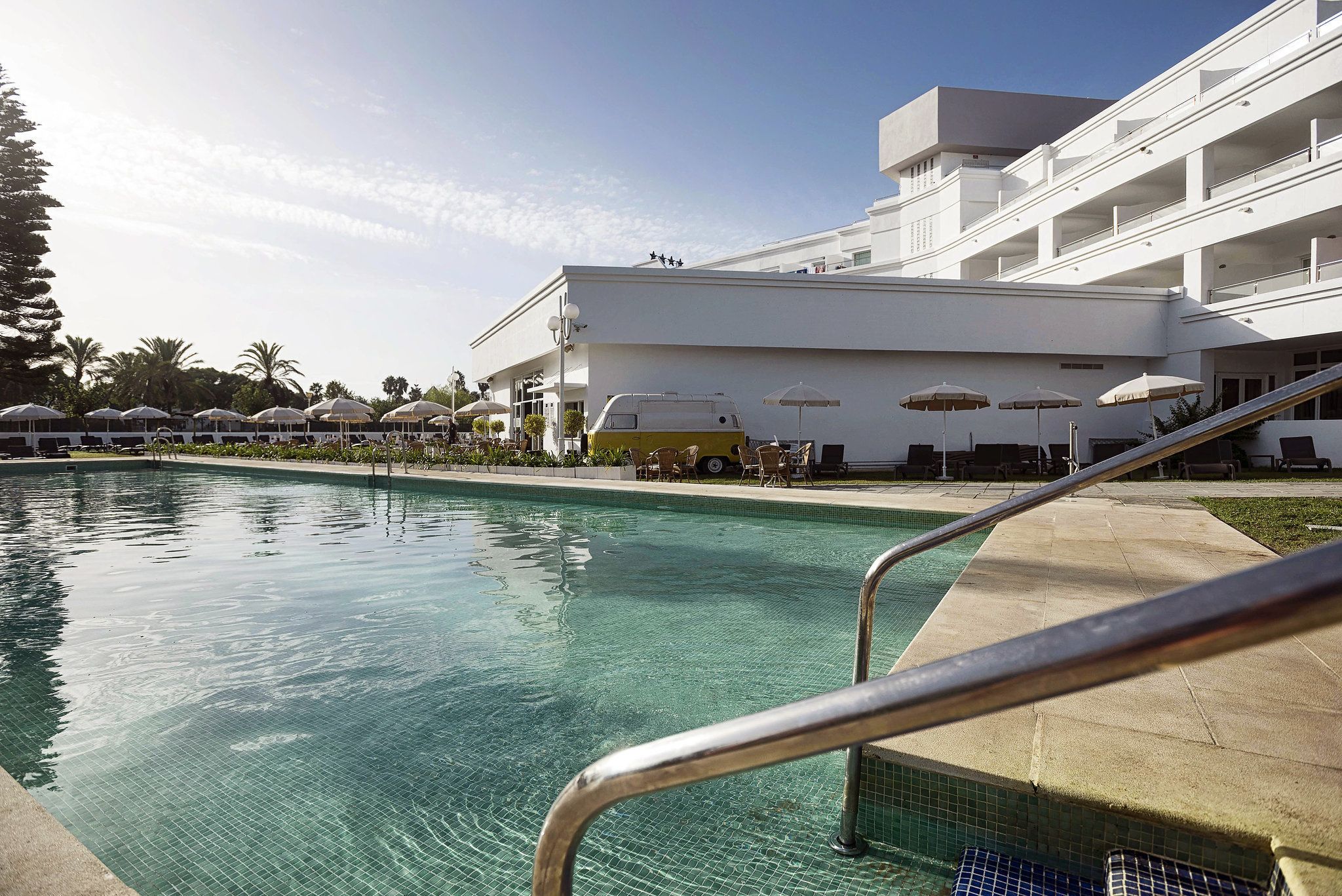Una piscina con superficie de agua azul cristalina, rodeada por un edificio blanco con varios balcones y sombrillas de exterior.