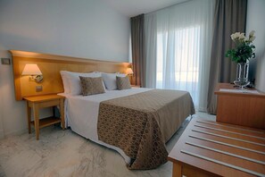 A hotel room with a large bed, two bedside tables with lamps, a wooden headboard, a bedside table with a vase of flowers, and a view of the outdoors through a window with curtains. - ALEGRIA Costa Ballena (Rota)