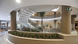 A modern indoor atrium with a central staircase, a palm tree, and a seating area with chairs. - ALEGRIA Costa Ballena (Rota)