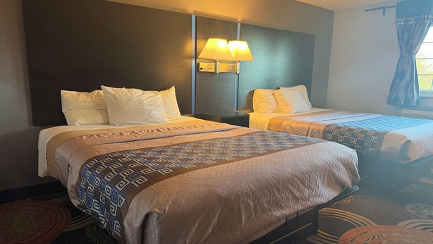 A hotel room with two beds, a nightstand with a lamp, and a window with curtains. -  Coratel Inn and Suites by Jasper Stillwater (Stillwater)